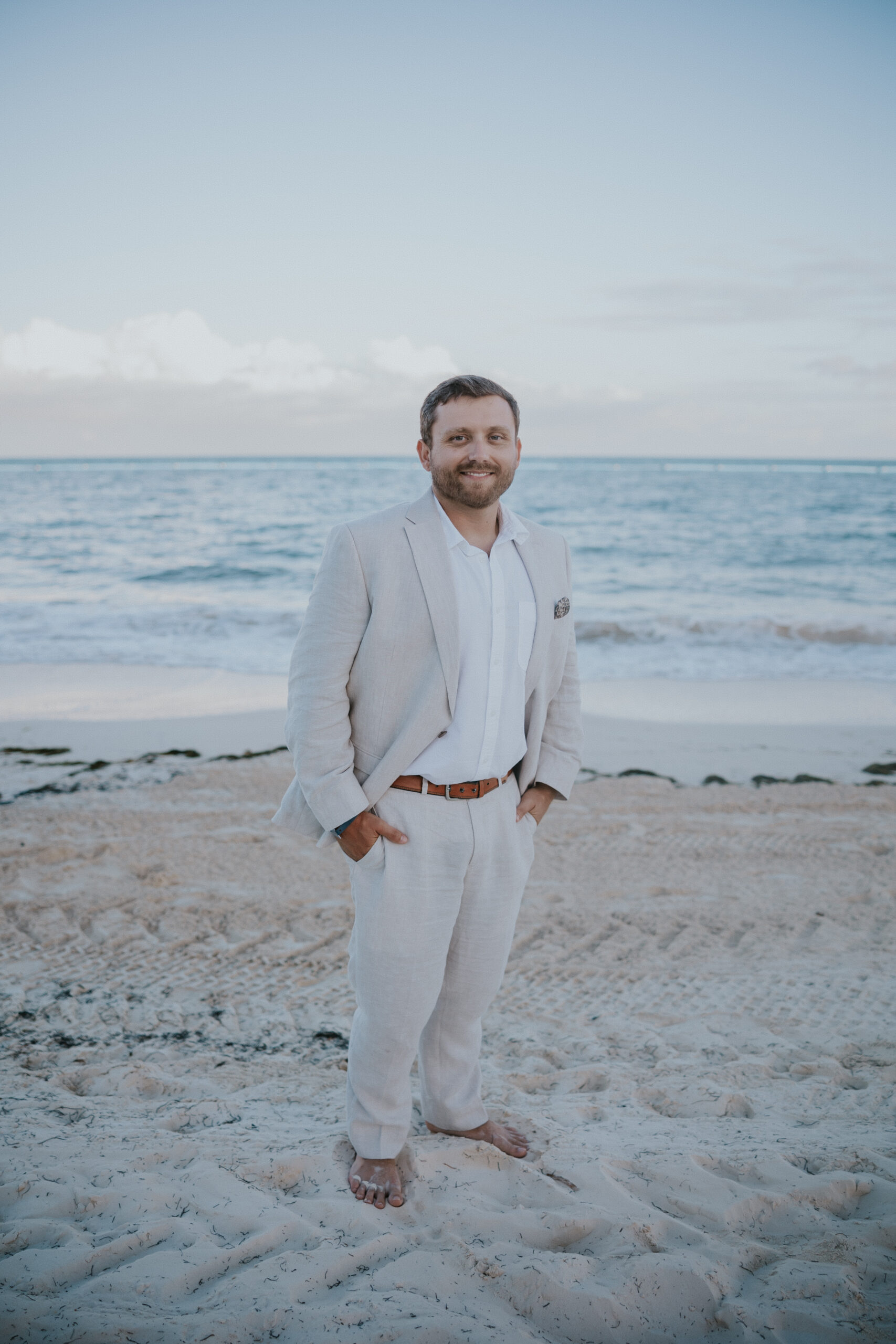 Groom standing barefoot on the sand in a light linen suit, smiling with the ocean behind him.
