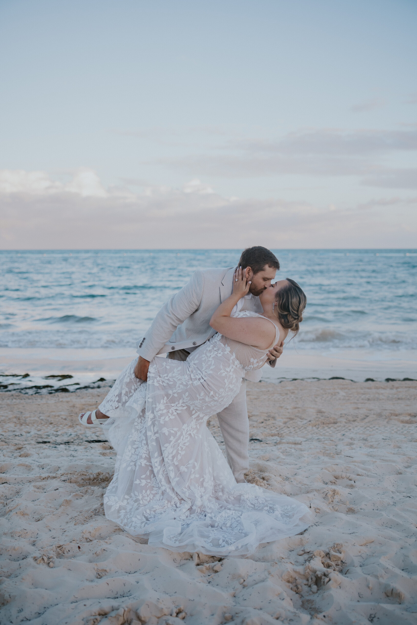 Groom dips the bride for a kiss in the sand, framed by soft sunset light over the ocean.
