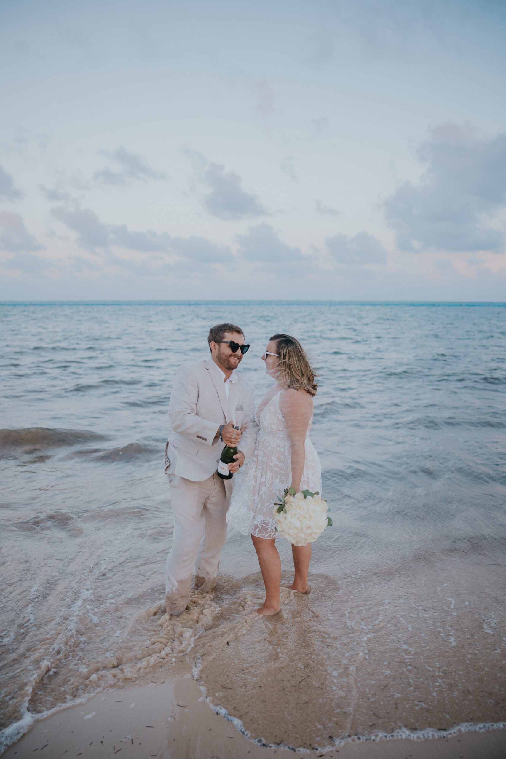 The couple shares a quiet moment, standing barefoot in the surf with the ocean behind them.