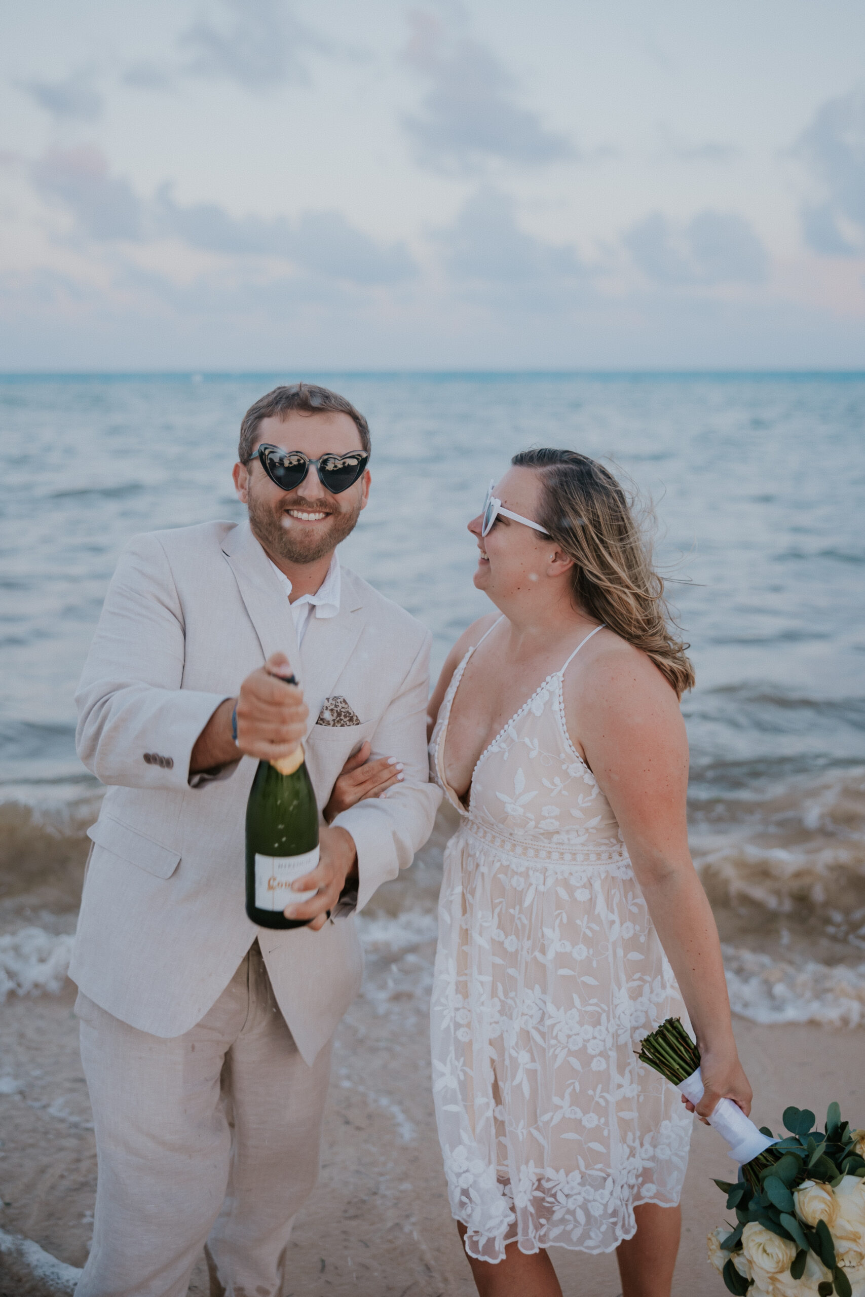 Newlyweds laugh together in sunglasses as the groom opens a bottle of champagne on the beach at their Mexico destination wedding.
