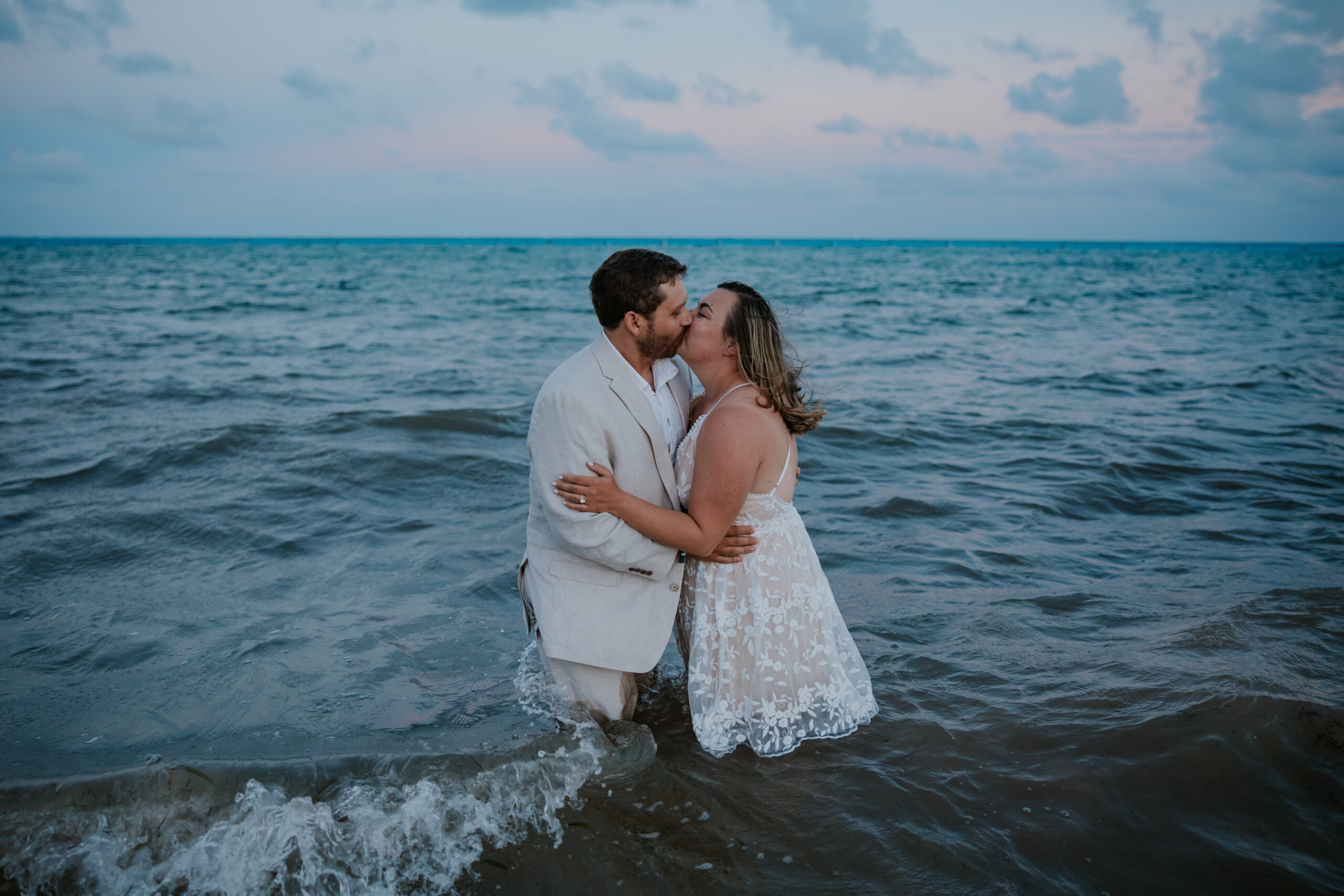 Couple wades into the sea at sunset, kissing in the waves in a joyful post-ceremony moment.