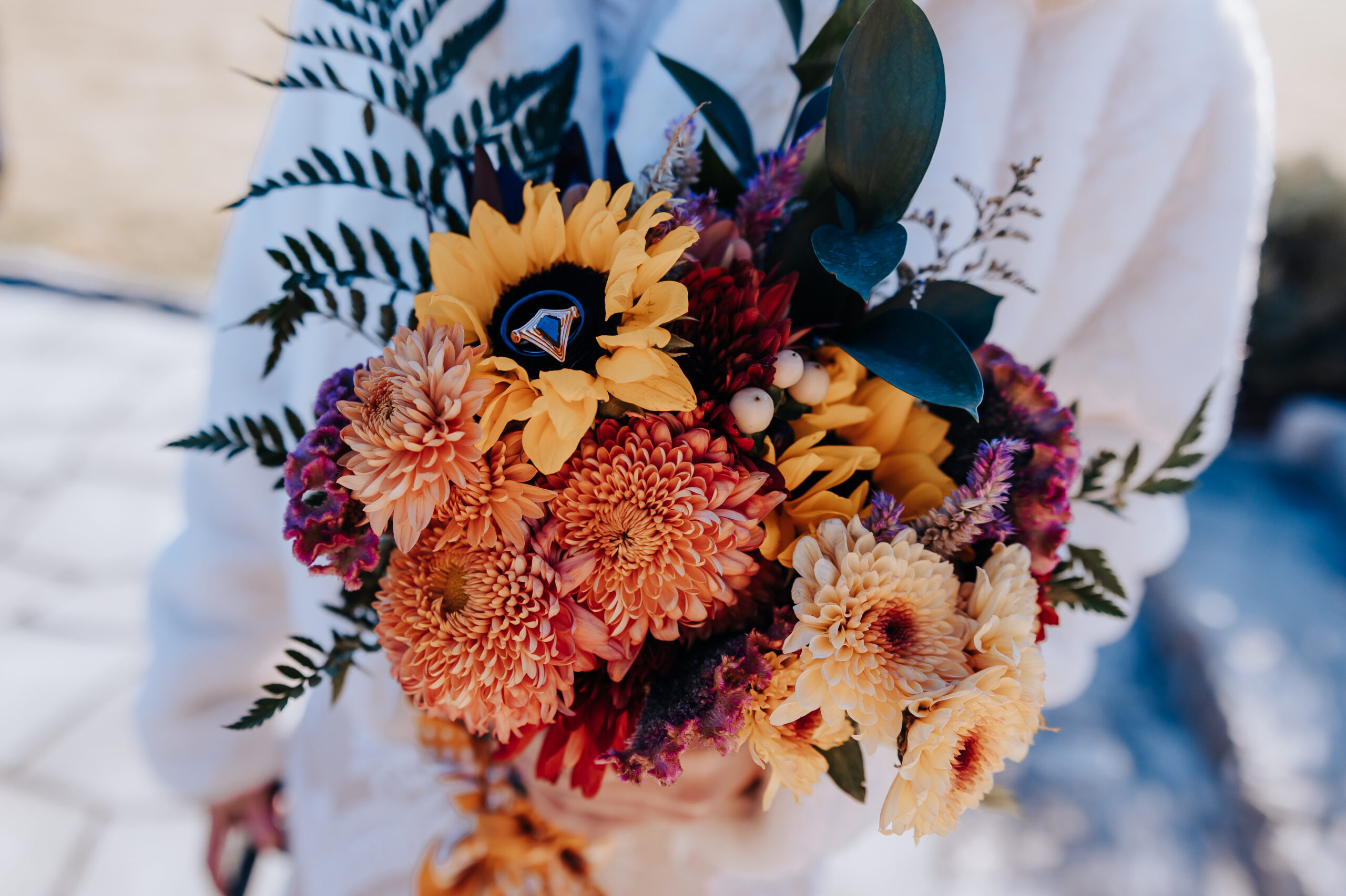 Bright autumn bouquet with wedding rings tucked into the sunflowers, held by the bride in a cozy white coat.