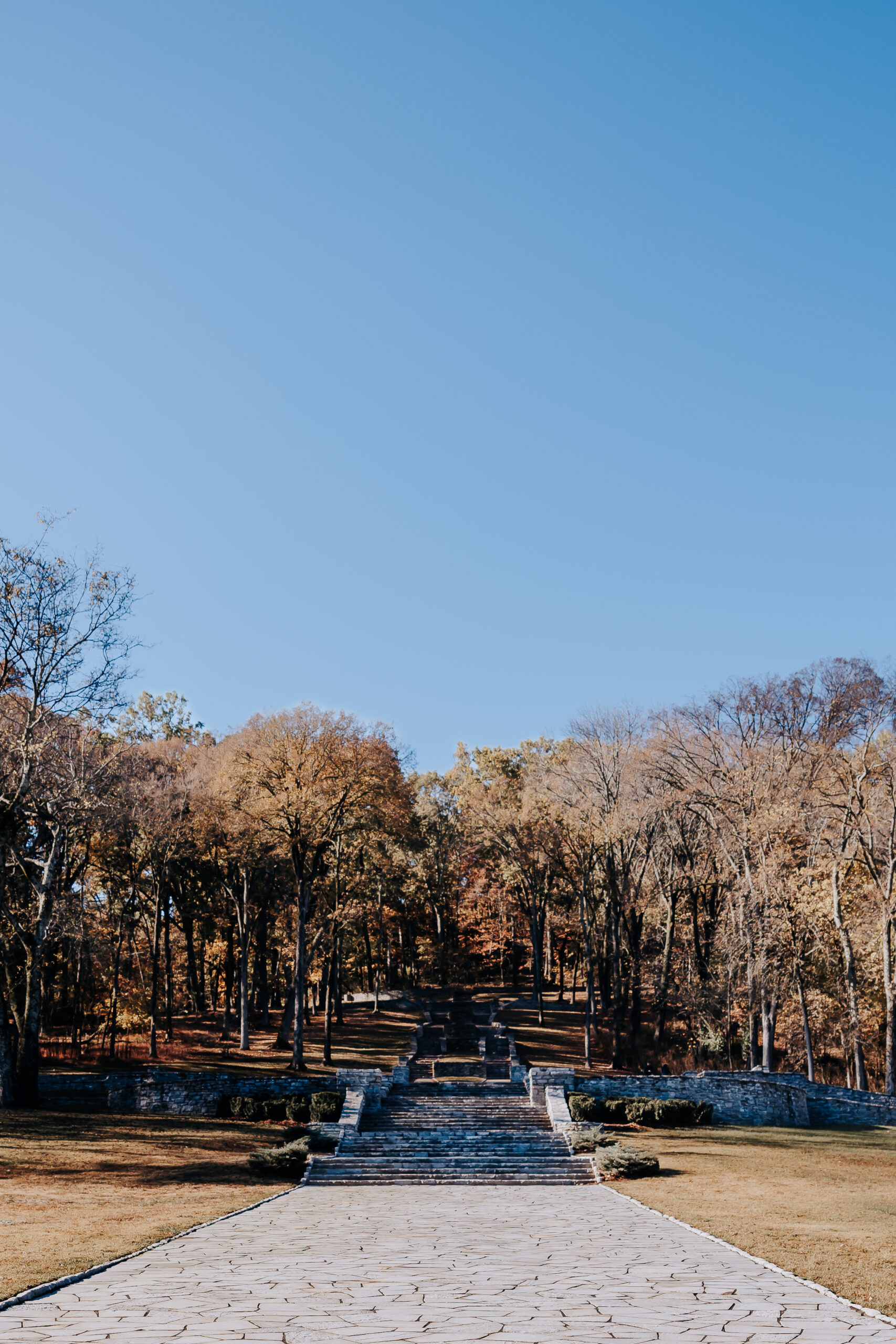 Scenic view of a long stone walkway and staircase framed by towering autumn trees under a clear blue sky.