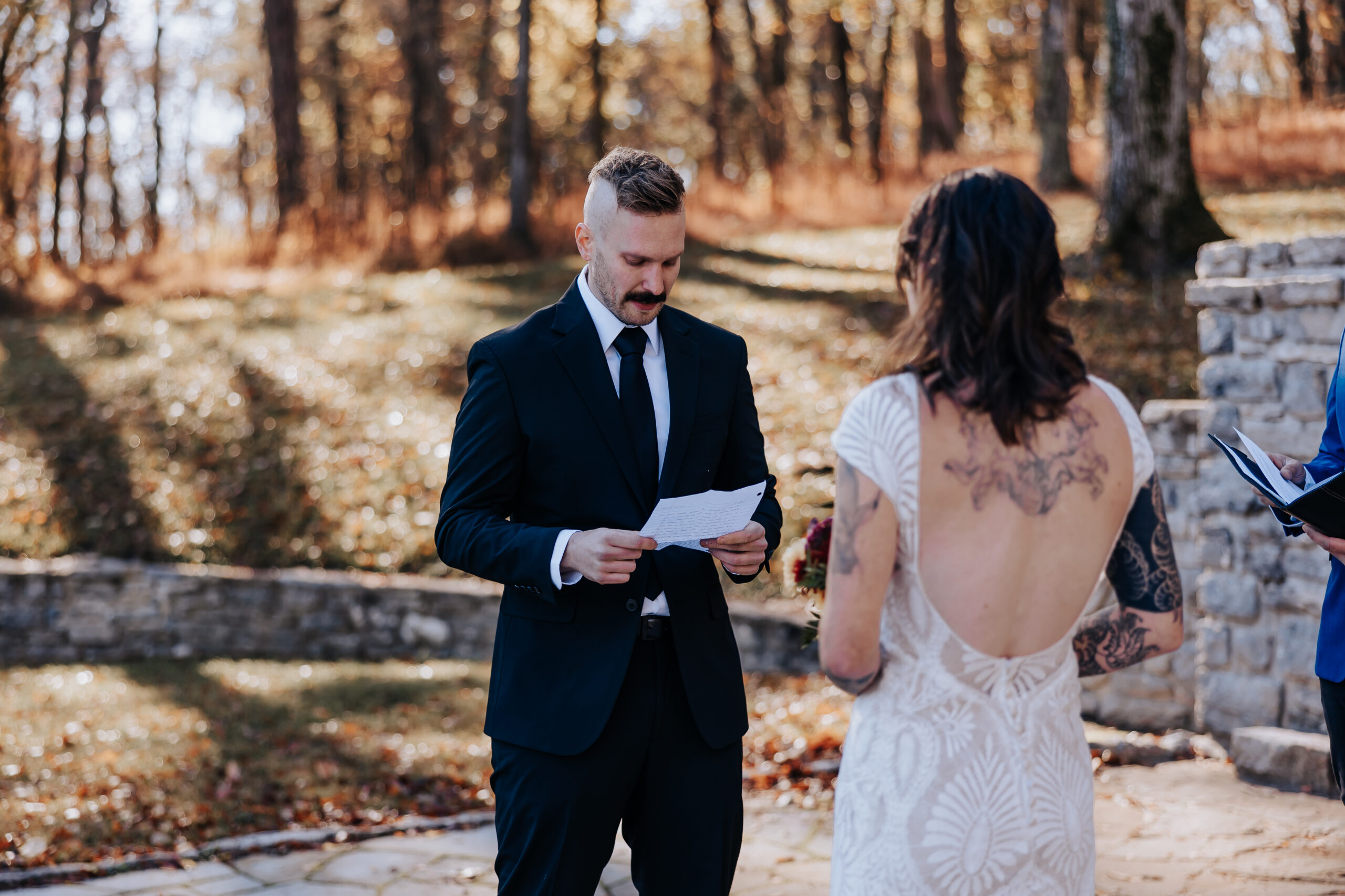 Groom reading heartfelt vows to the bride during an emotional outdoor ceremony surrounded by fall trees.