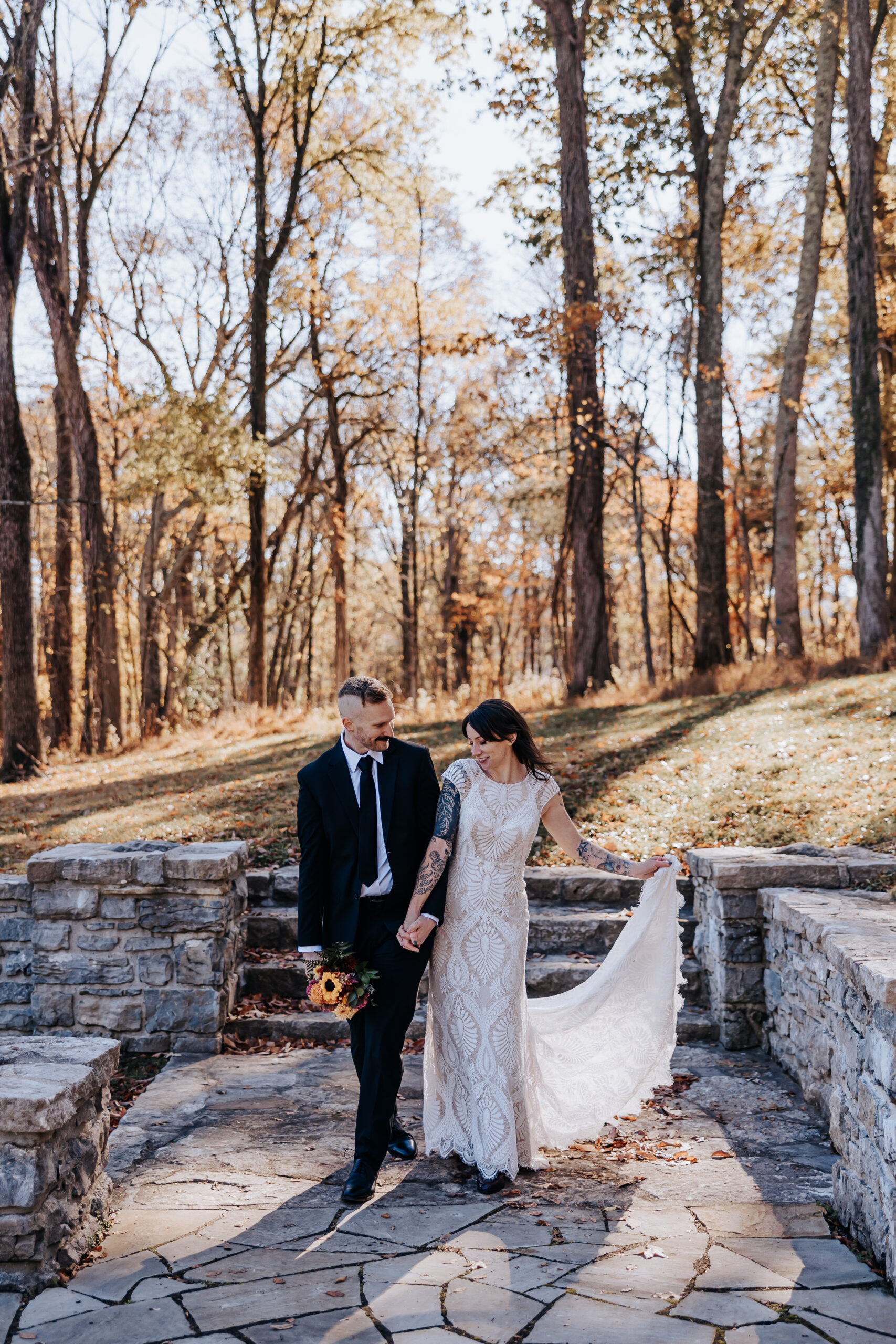 Couple strolls through a wooded stone walkway, bride lifting her dress with one hand and bouquet in the other at their wedding in Nashville.