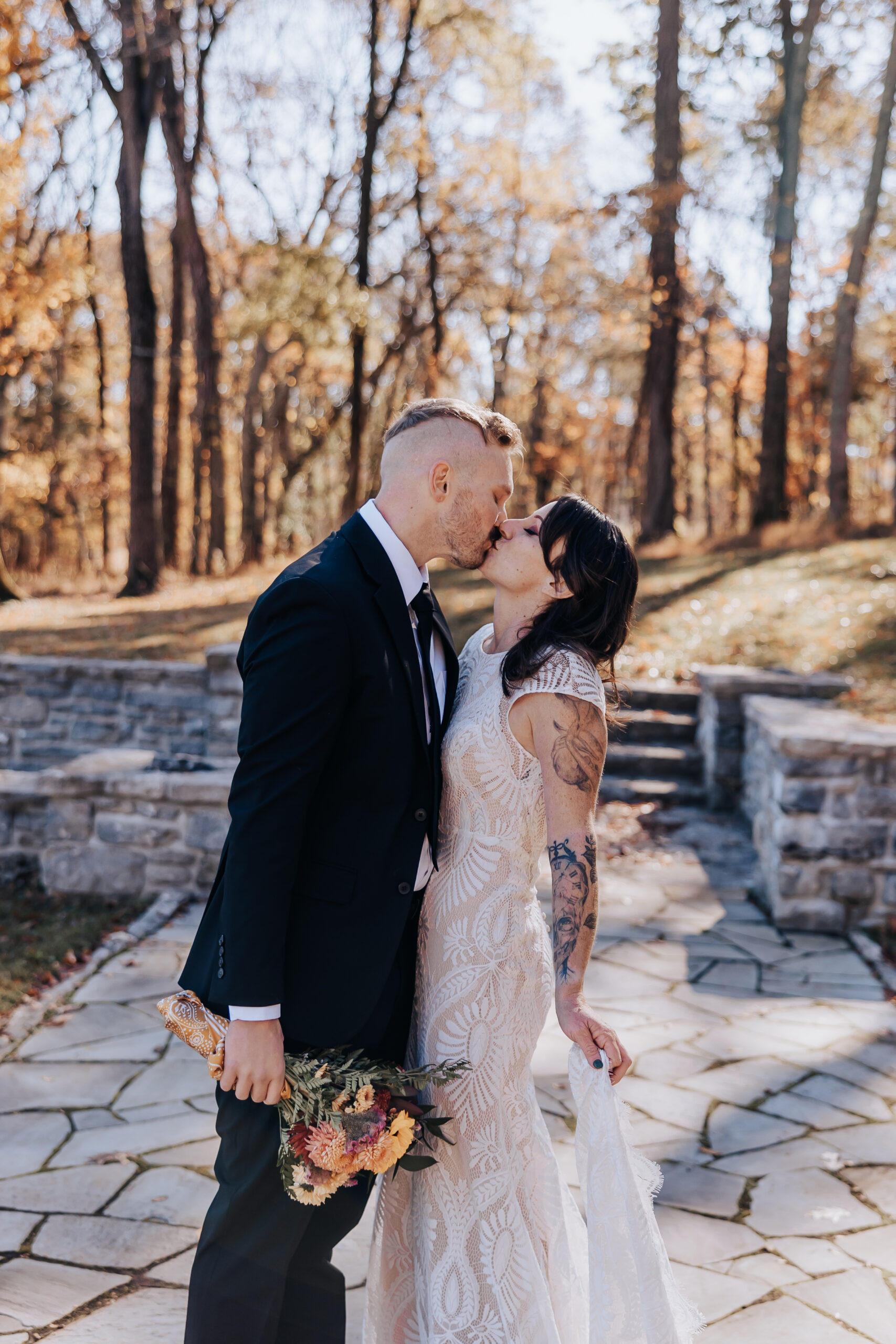 Bride and groom share a romantic kiss while holding a rustic bouquet in a golden forest setting.