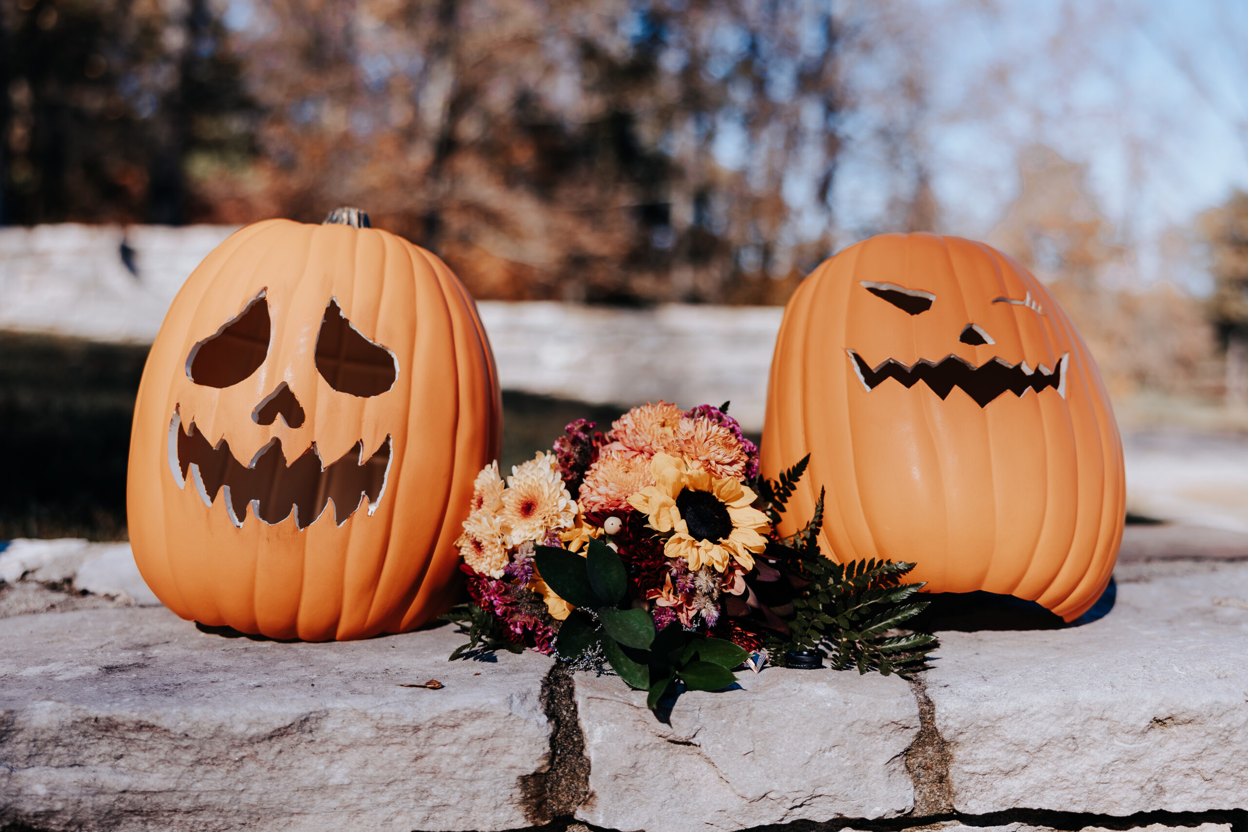 Two carved pumpkins sit on a stone ledge next to a colorful bouquet of sunflowers and mums, adding whimsy to this fall wedding scene.