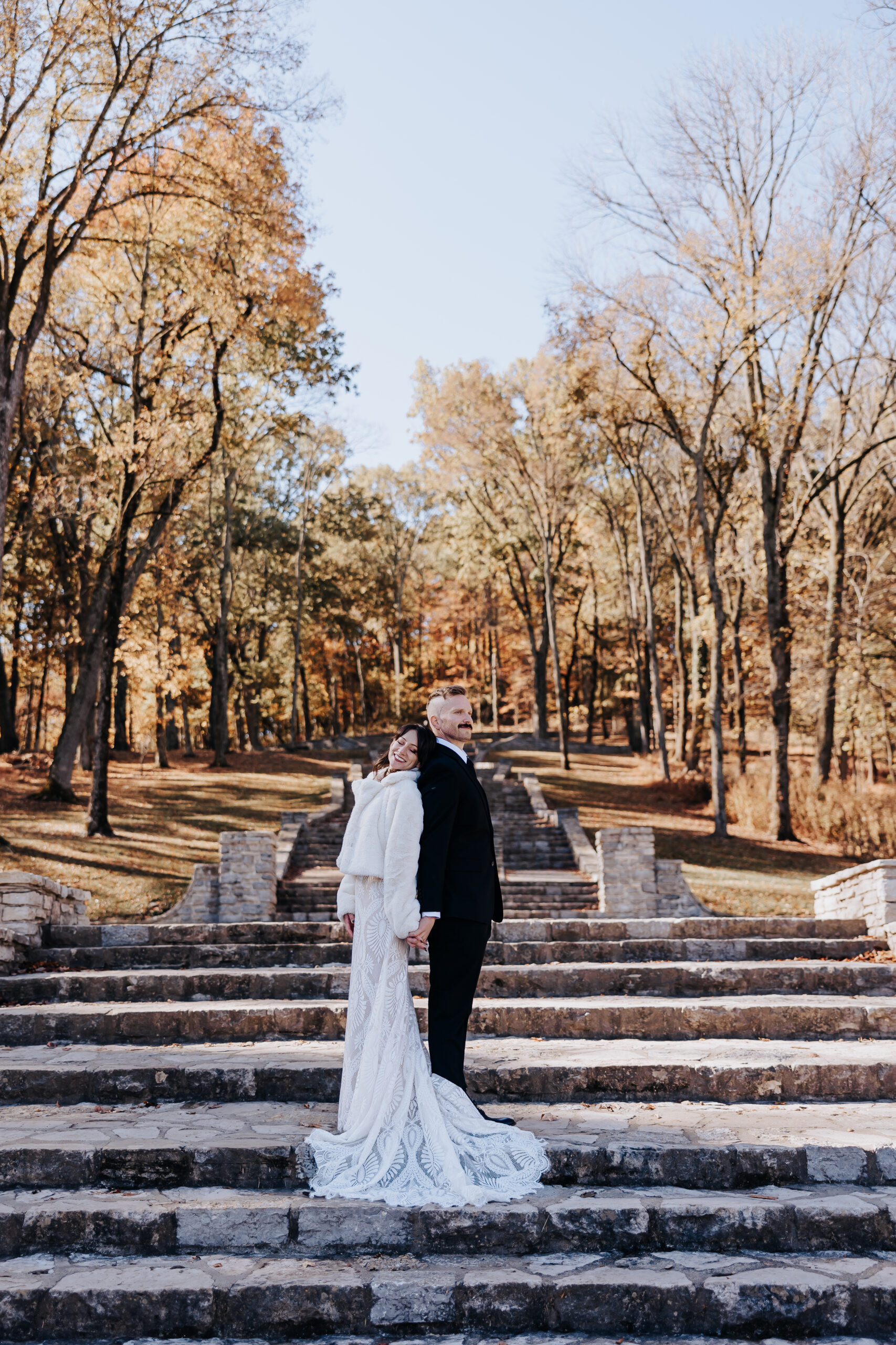 Newlyweds stand back-to-back on the stone staircase framed by tall trees — a quiet moment of connection during their fall wedding in Nashville.