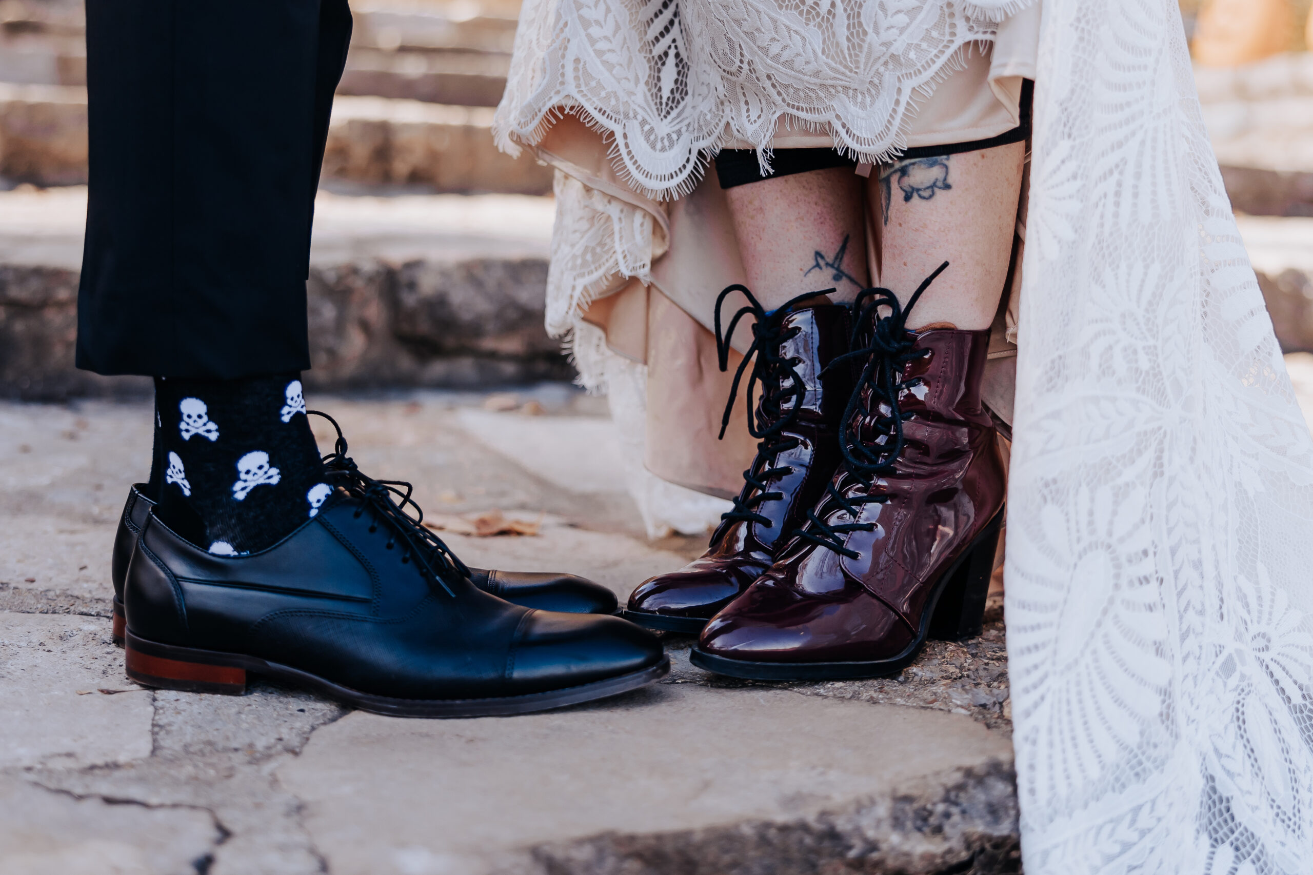 Close-up of the couple’s unique footwear: shiny burgundy boots and skull socks peeking from under a lace wedding dress.