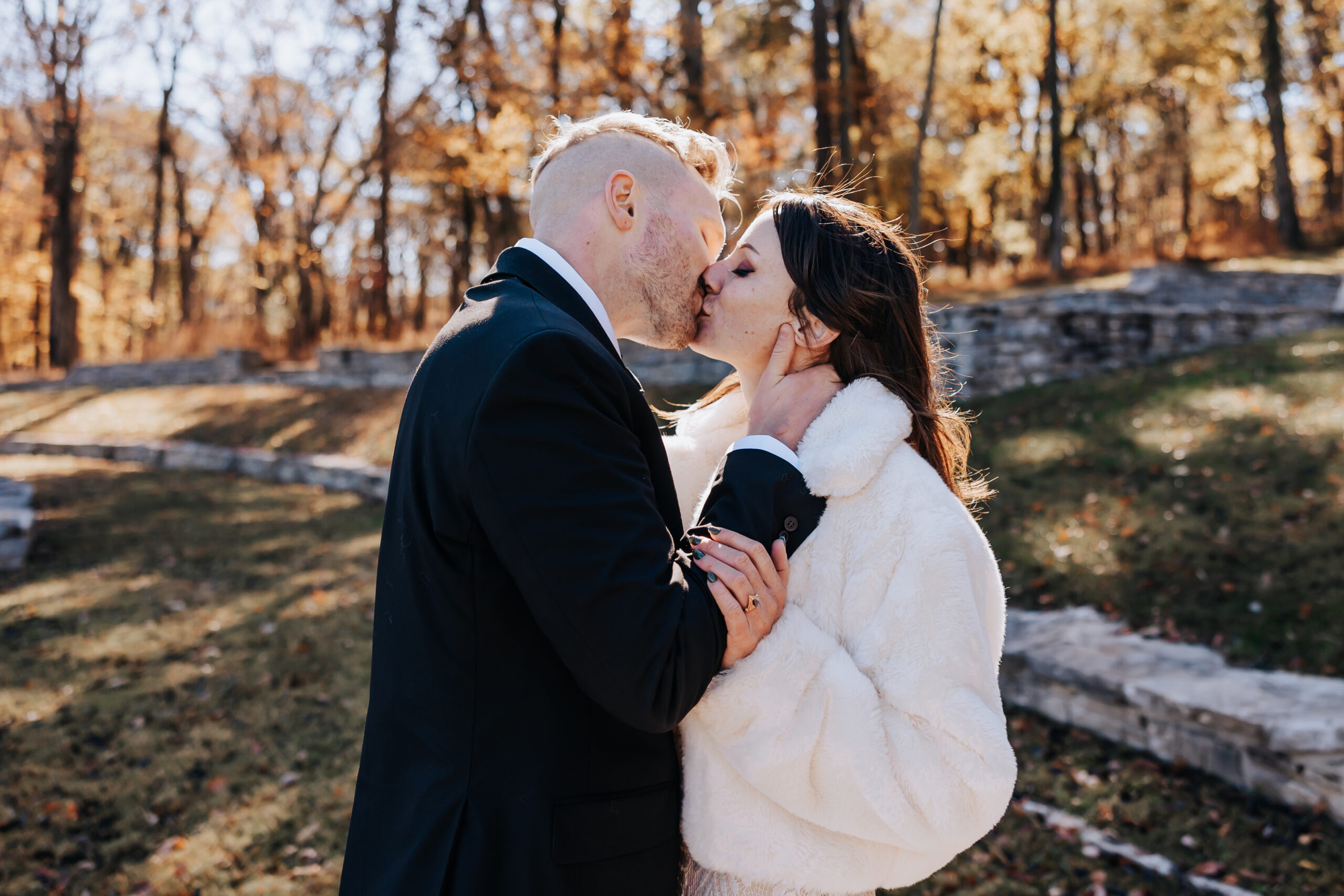 Sweet moment as the couple shares a kiss outdoors, the bride wrapped in a cozy white shawl on a crisp fall day.