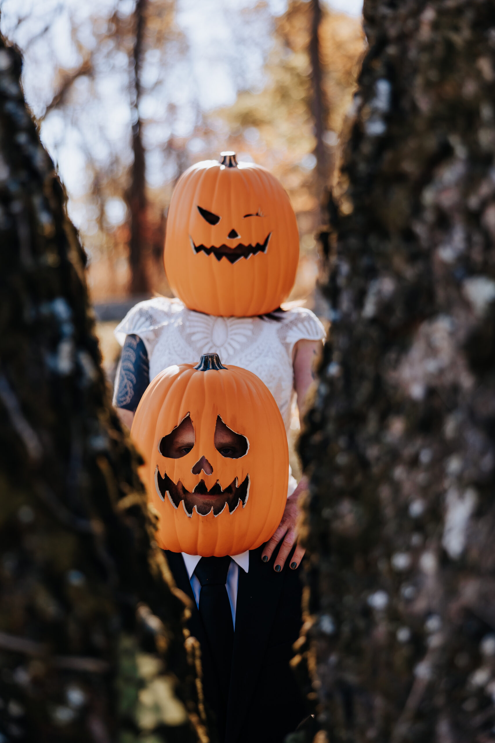 Playful couple wearing carved pumpkin masks peeks through tree trunks in a quirky Halloween-themed wedding in Nashville.