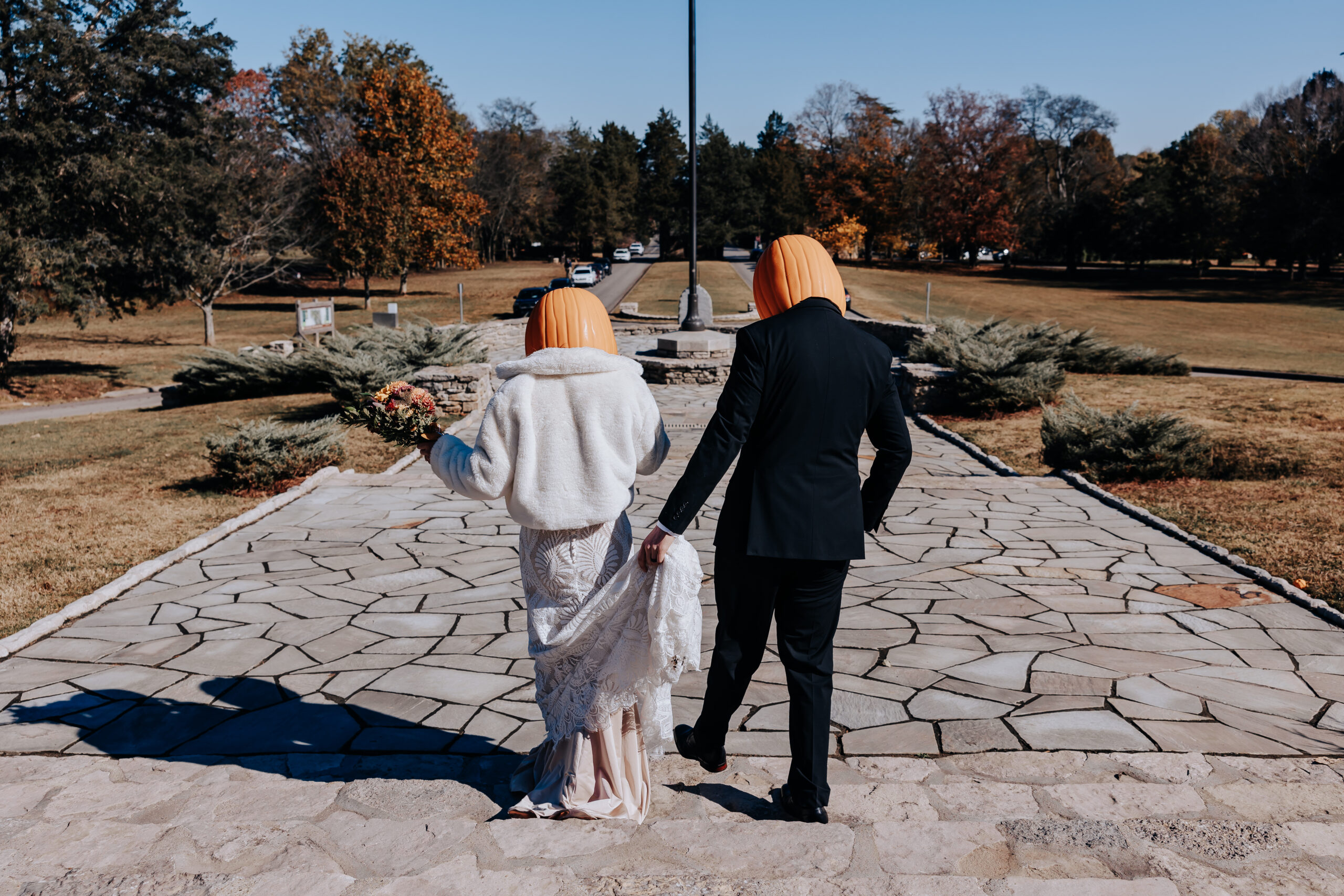 Couple walking hand-in-hand down a stone path wearing pumpkin masks — a fun twist on tradition.
