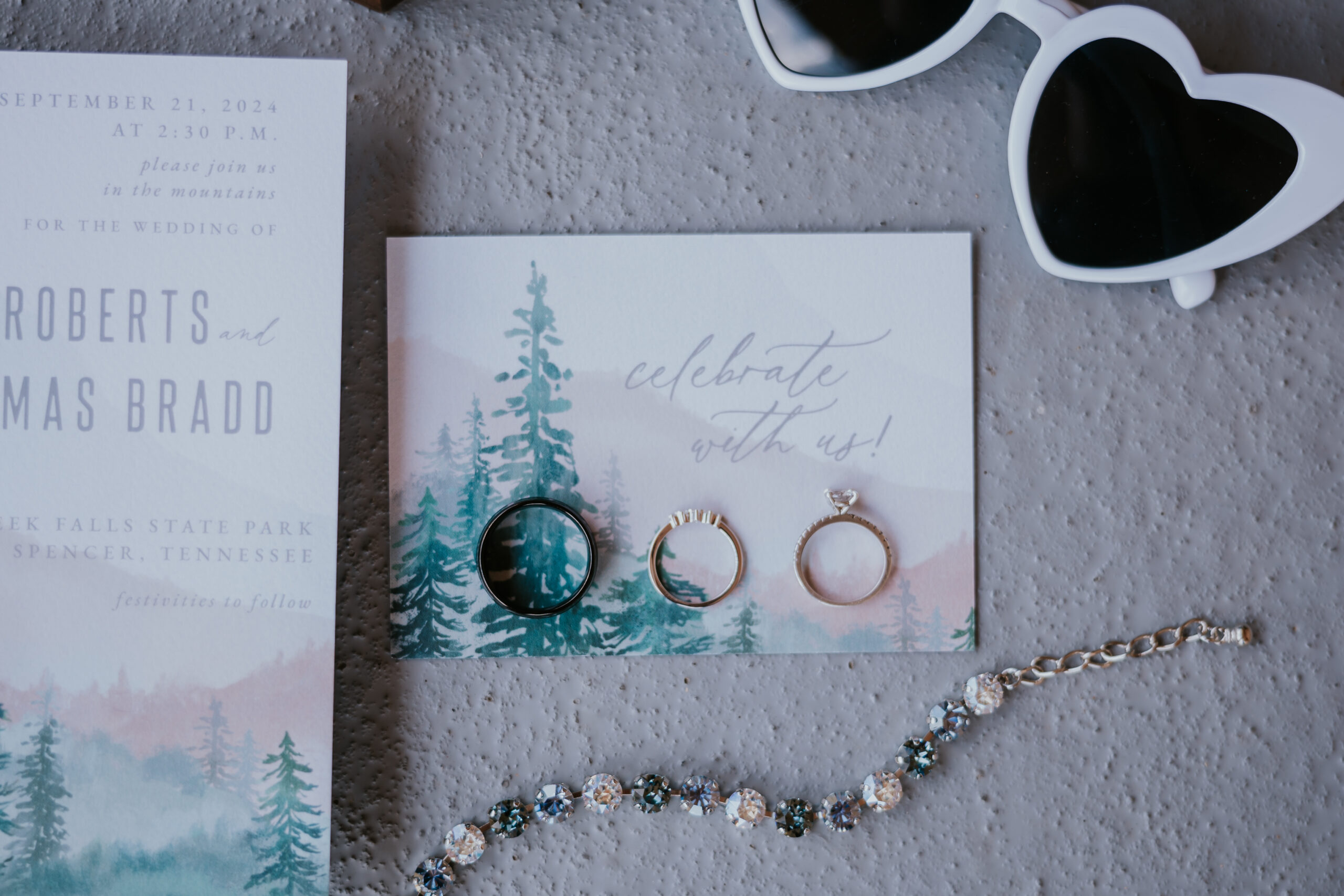 Flat lay of wedding invitation, rings, and accessories with heart-shaped sunglasses and woodland-themed design, tying together rustic romance and modern flair.
