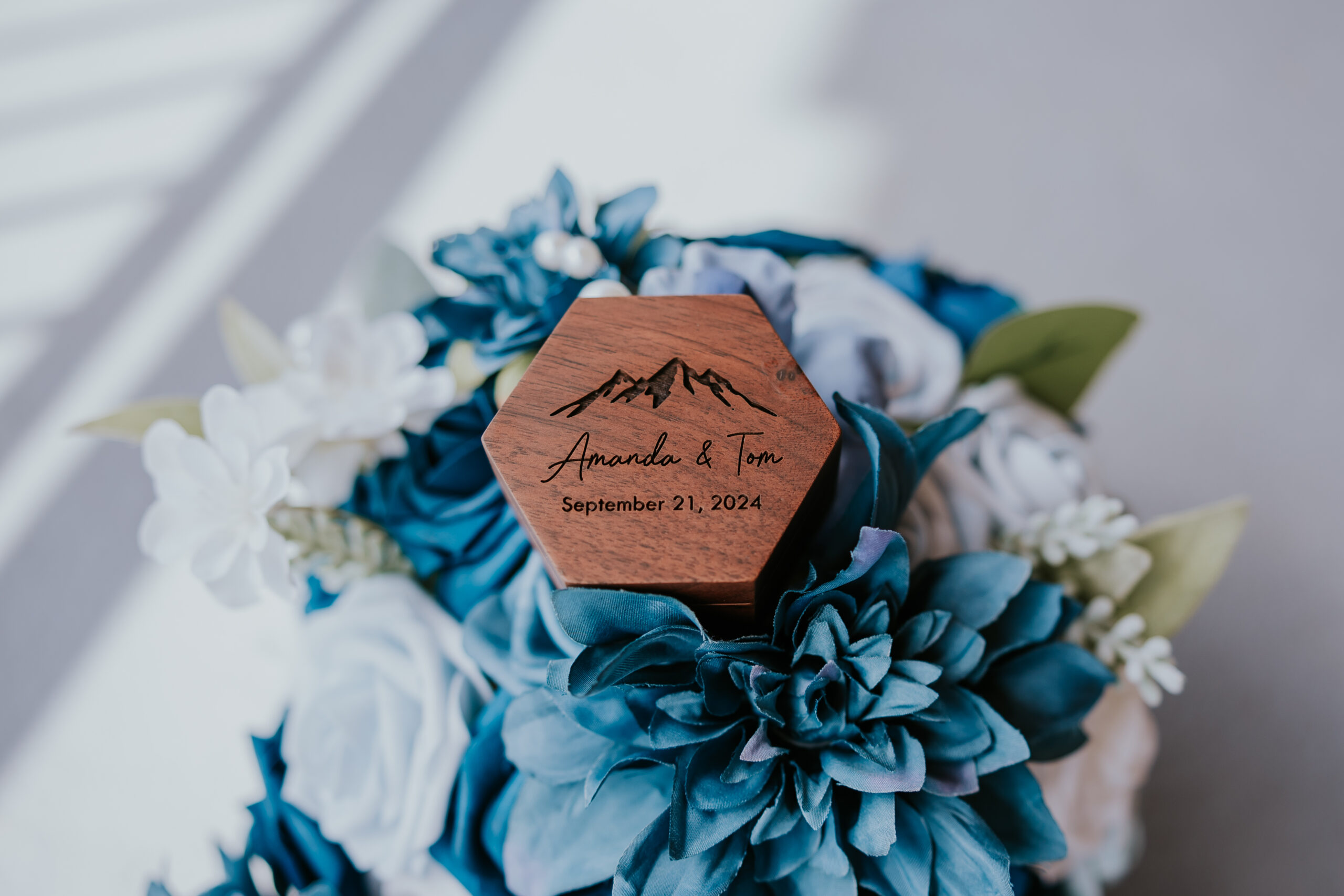 Custom hexagon wood tag reading “Amanda & Tom – September 21, 2024” nestled in a bouquet of blue and white flowers, a personalized touch that reflects unique wedding ideas.
