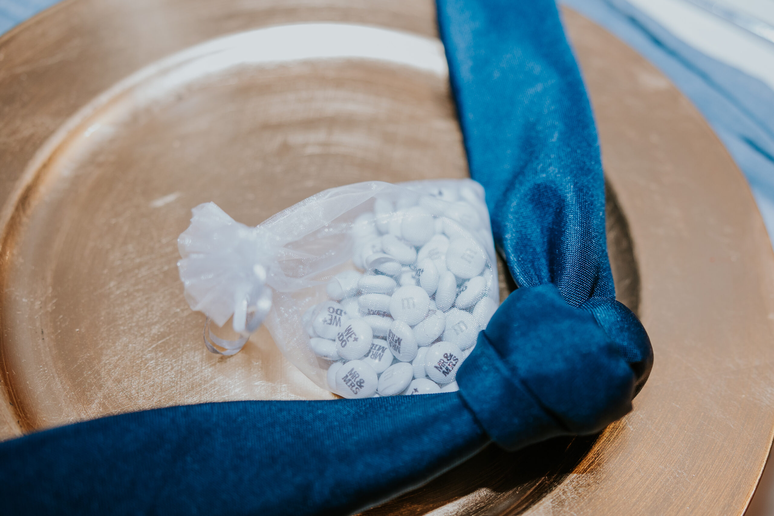 Close-up of a gold charger plate with navy napkin and a favor bag of custom wedding M&Ms — a fun detail showcasing unique wedding ideas for reception tables.