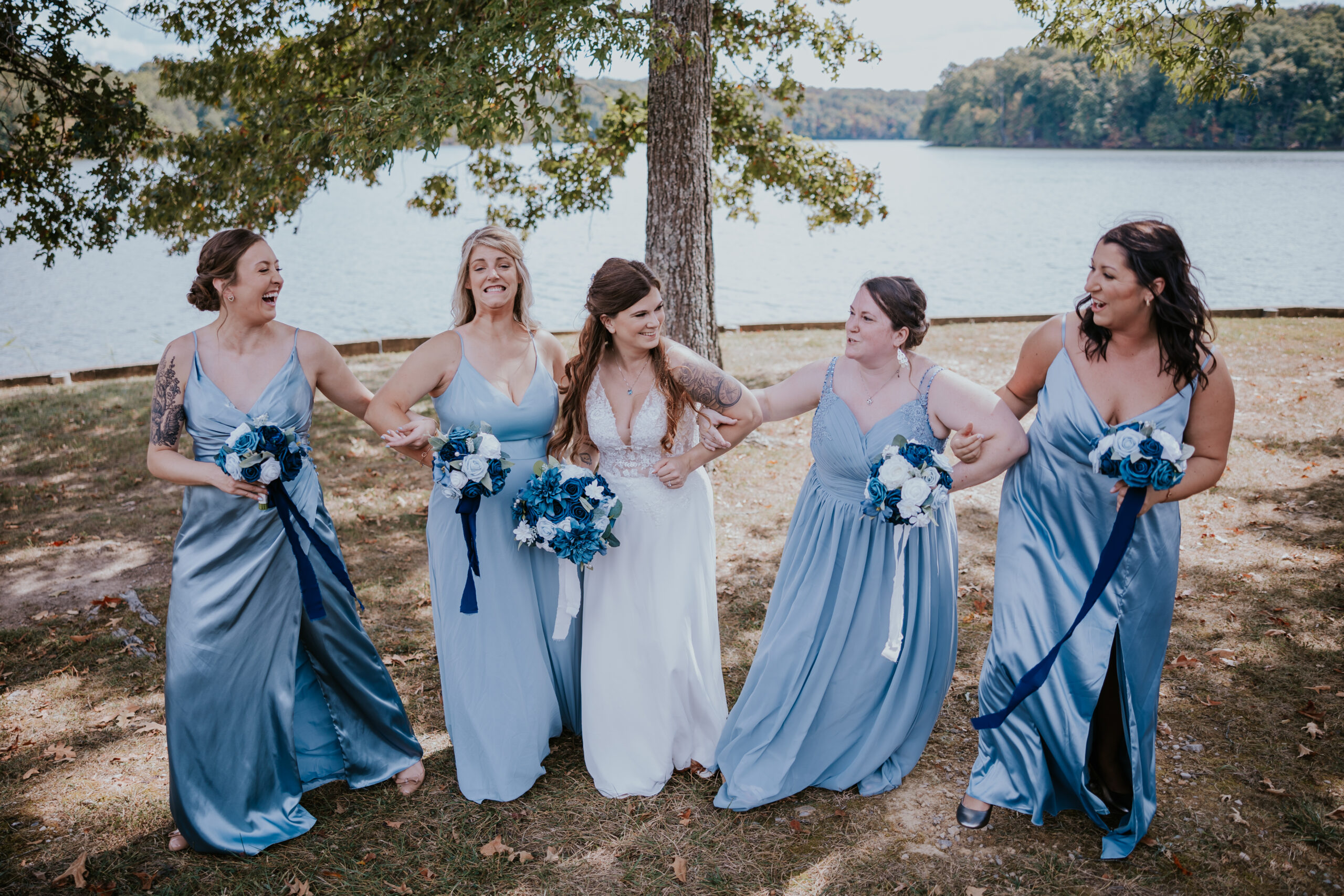 Bride and bridesmaids laughing by the lakeside in matching blue dresses, celebrating friendship and joyful wedding moments.