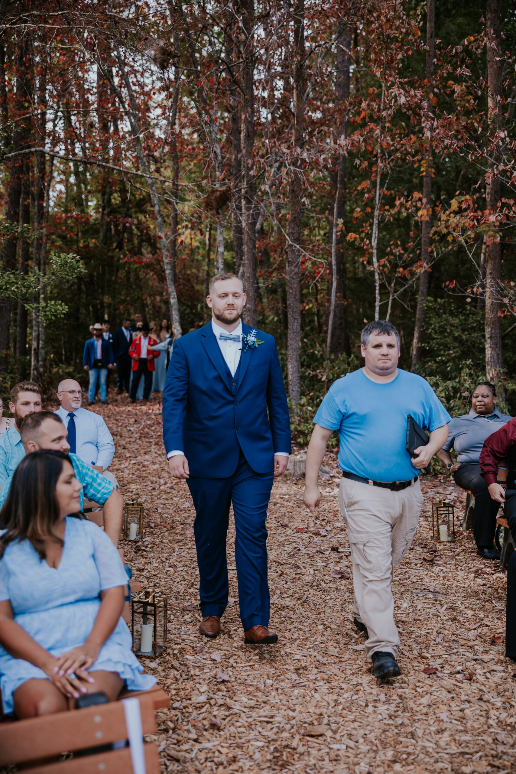 The groom walking down a rustic wooded aisle with a special guest, surrounded by fall foliage and supportive friends and family.
