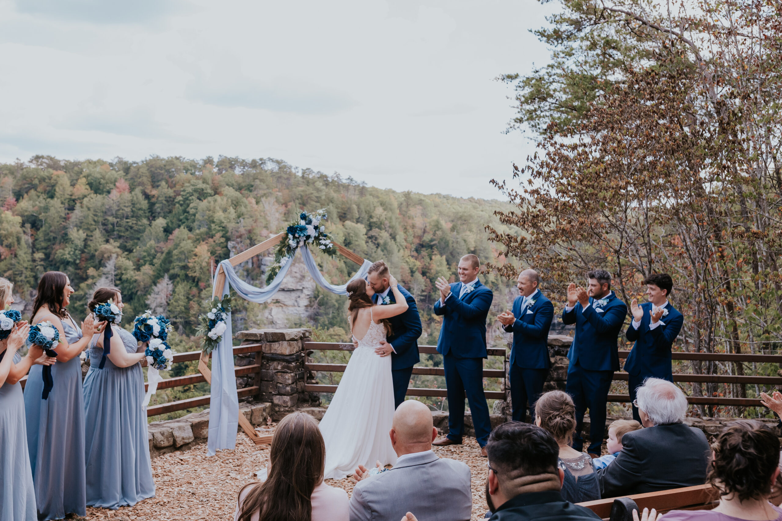 The bride and groom share their first kiss as a married couple, framed by cheering guests and the bridal party, with an epic Fall Creek Falls view in the background.
