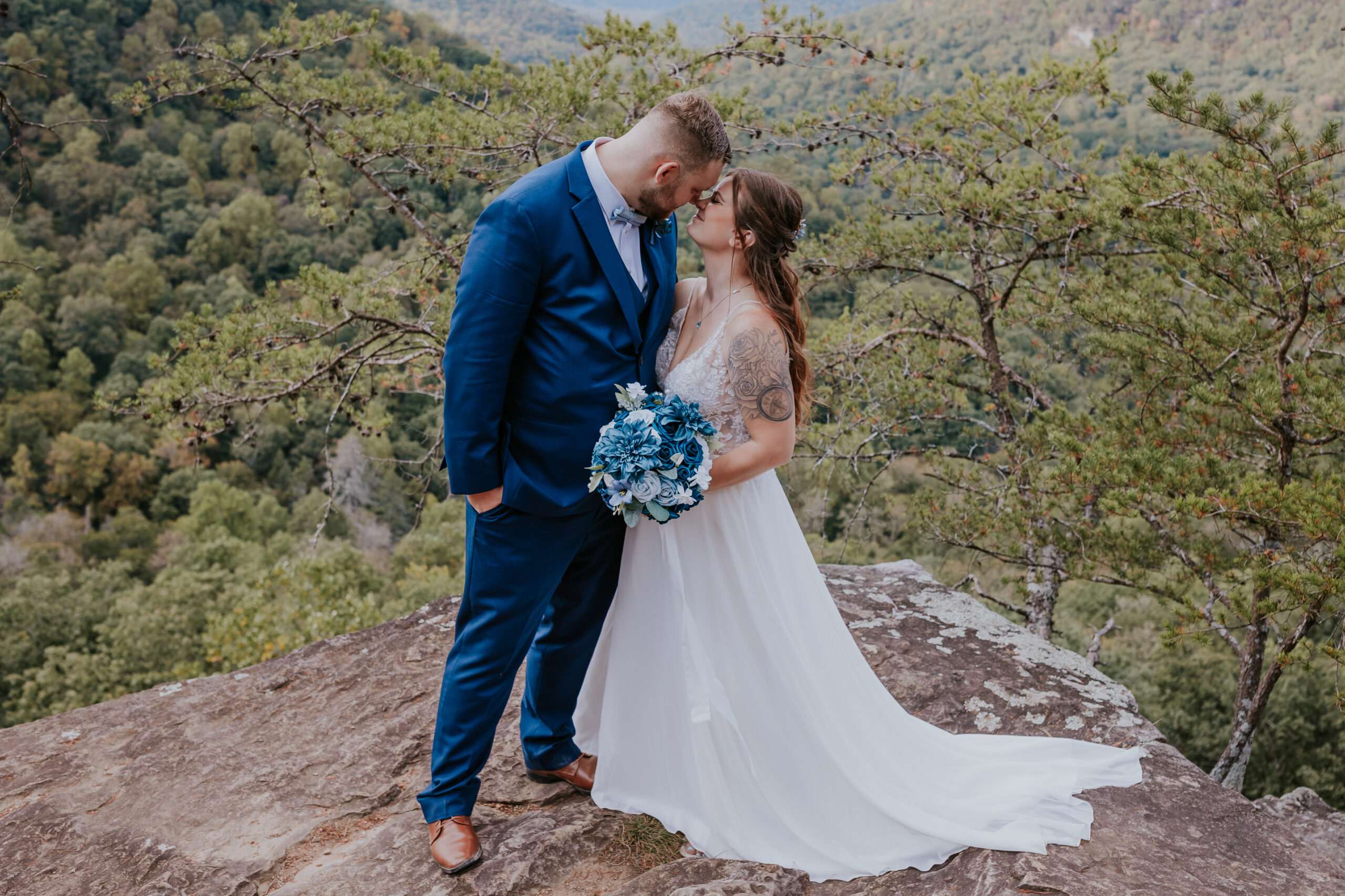 Bride and groom kiss on a rocky ledge, framed by a mountain backdrop and holding a bold blue bouquet — a striking example of unique wedding ideas for adventurous couples.