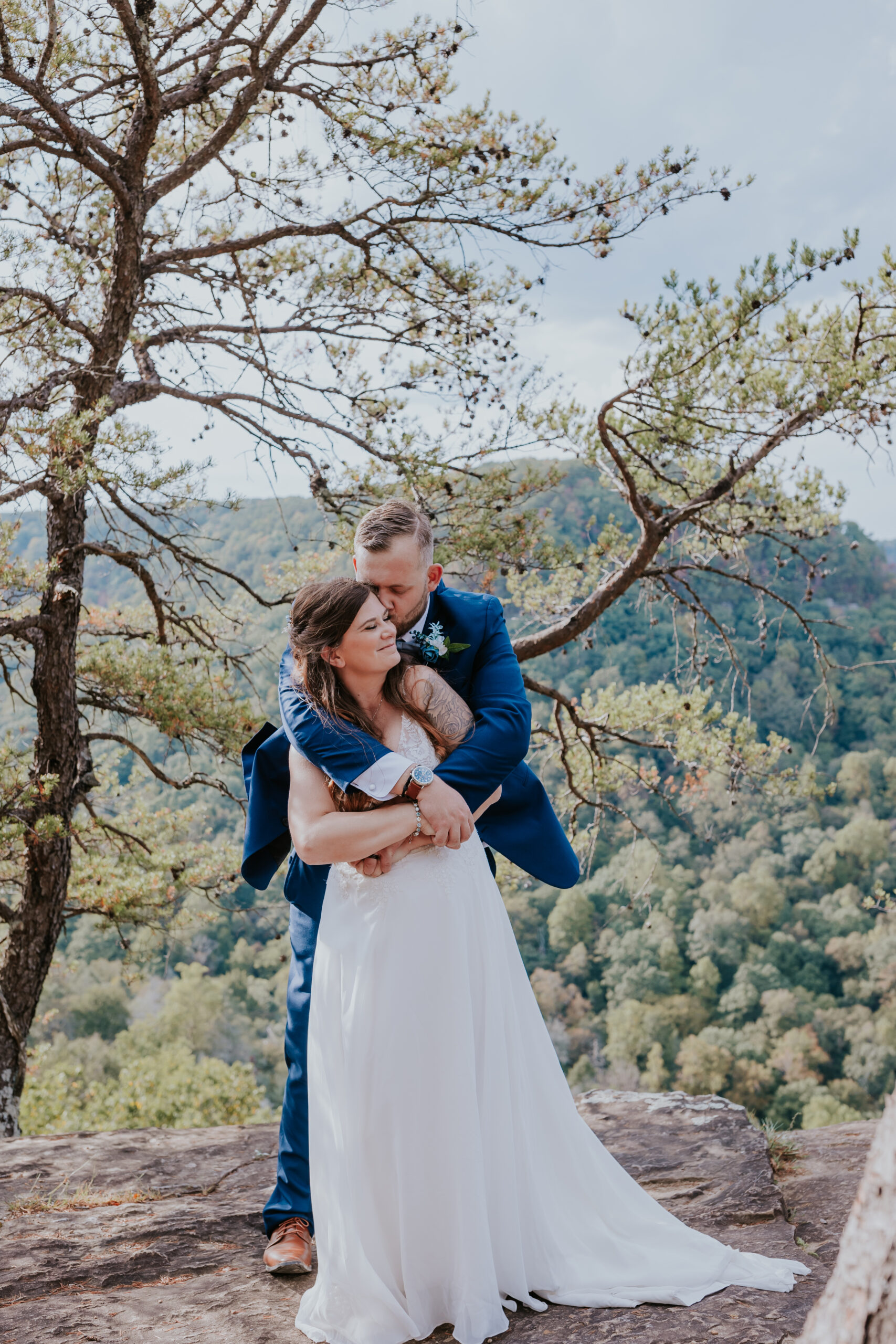 Groom wraps his arms around the bride as they stand on a rocky overlook, soaking in the peaceful forest views.