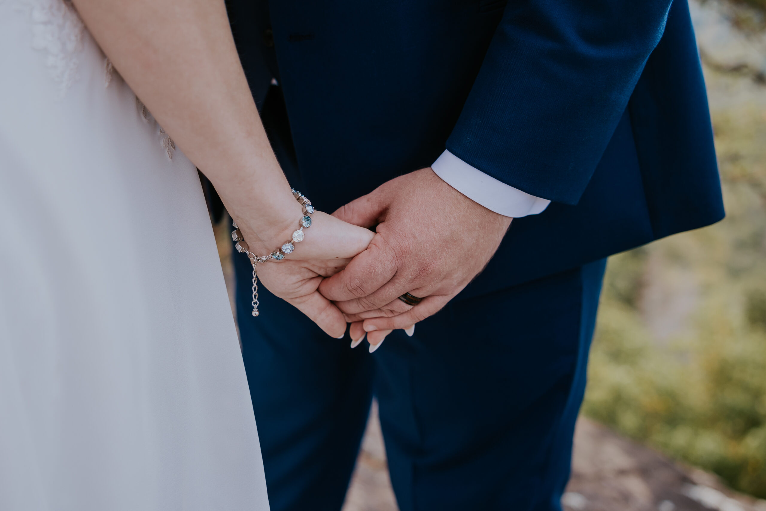 Bride and groom holding hands, featuring a sparkling bracelet and navy wedding suit, against a mountain overlook backdrop.