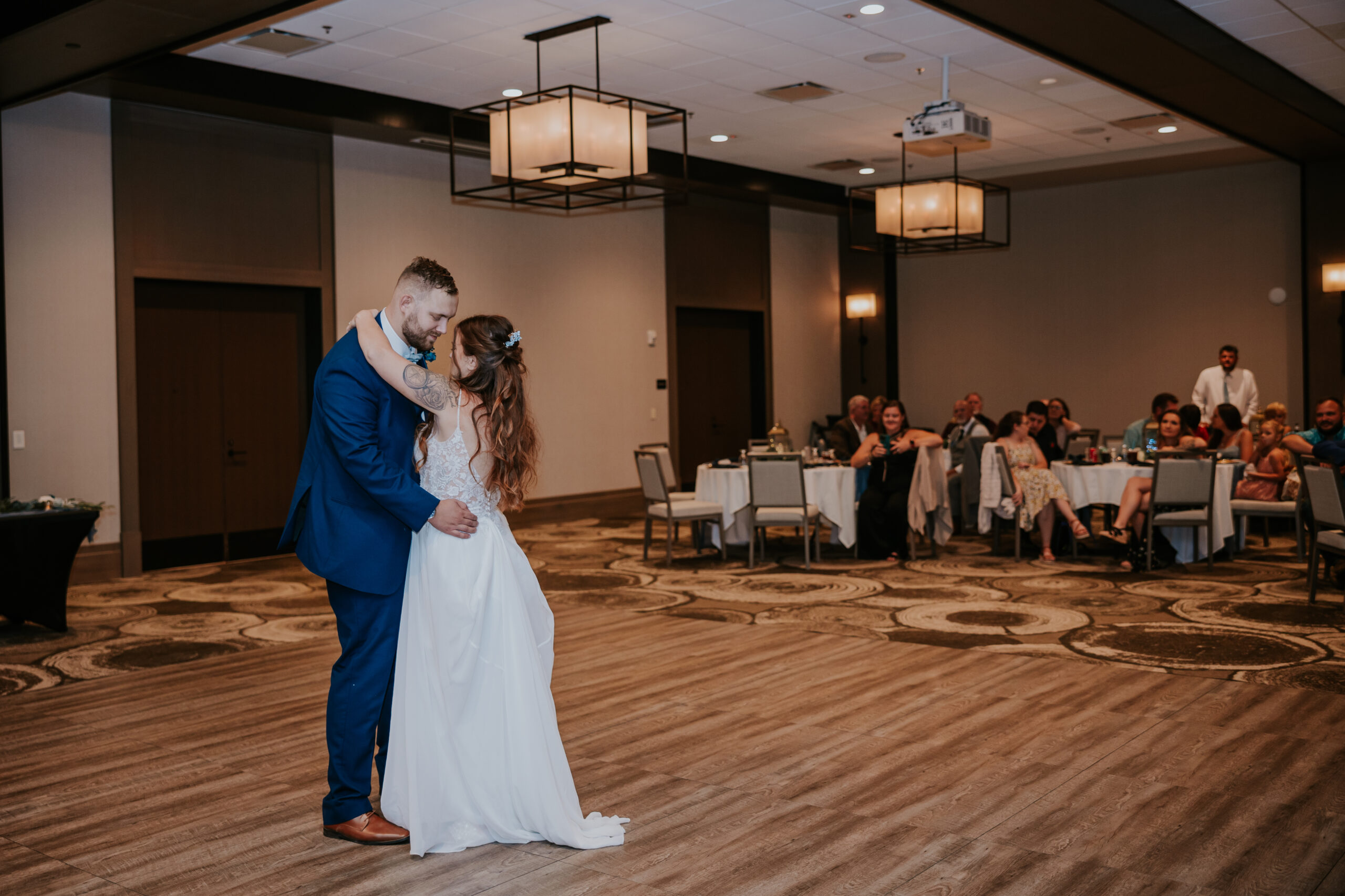 Bride and groom share their first dance in a warmly lit reception hall as guests look on with smiles.