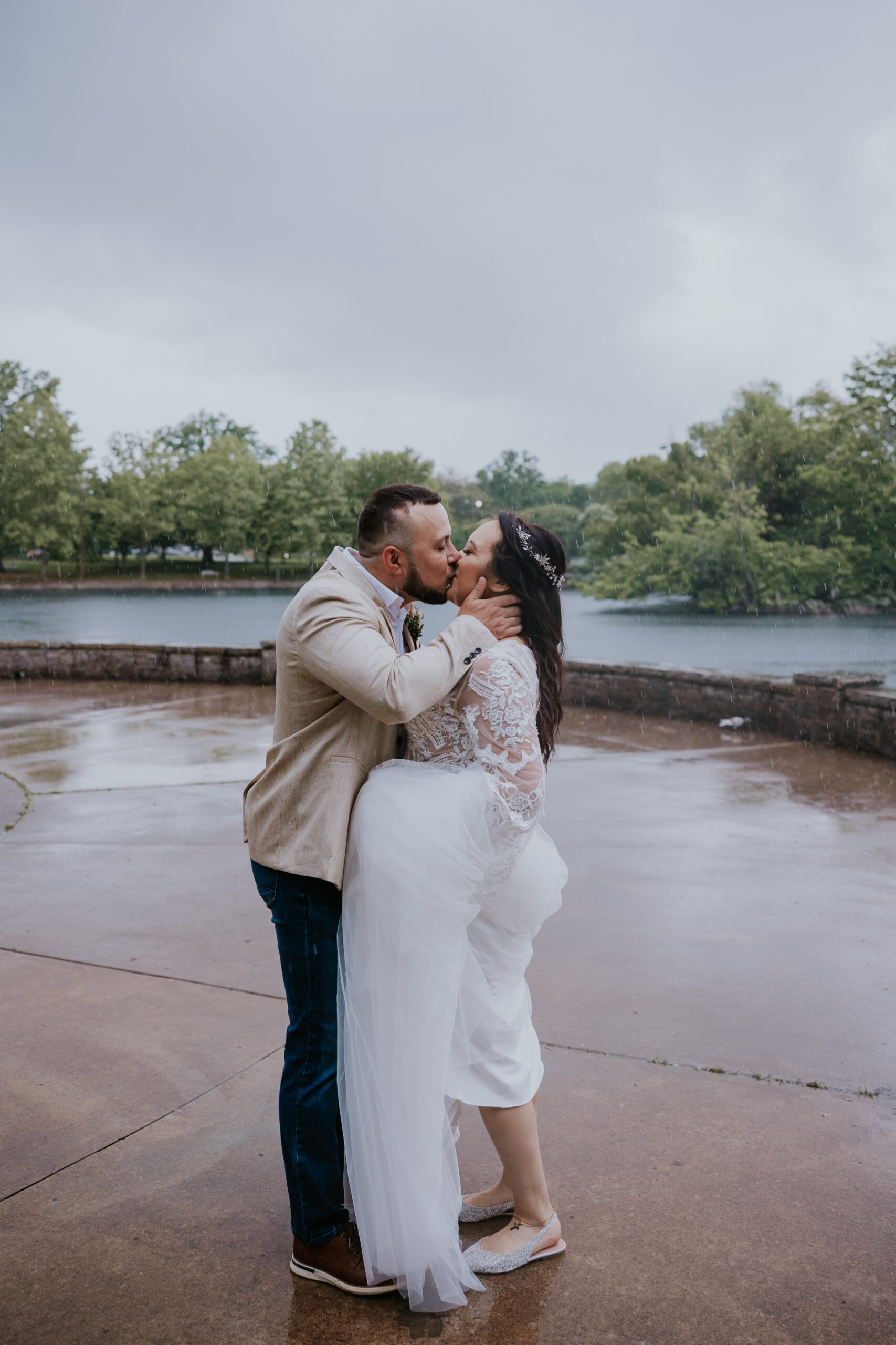 Rainy day kiss beside the water as groom lifts bride’s gown—an emotional moment beautifully captured by a travel wedding photographer.