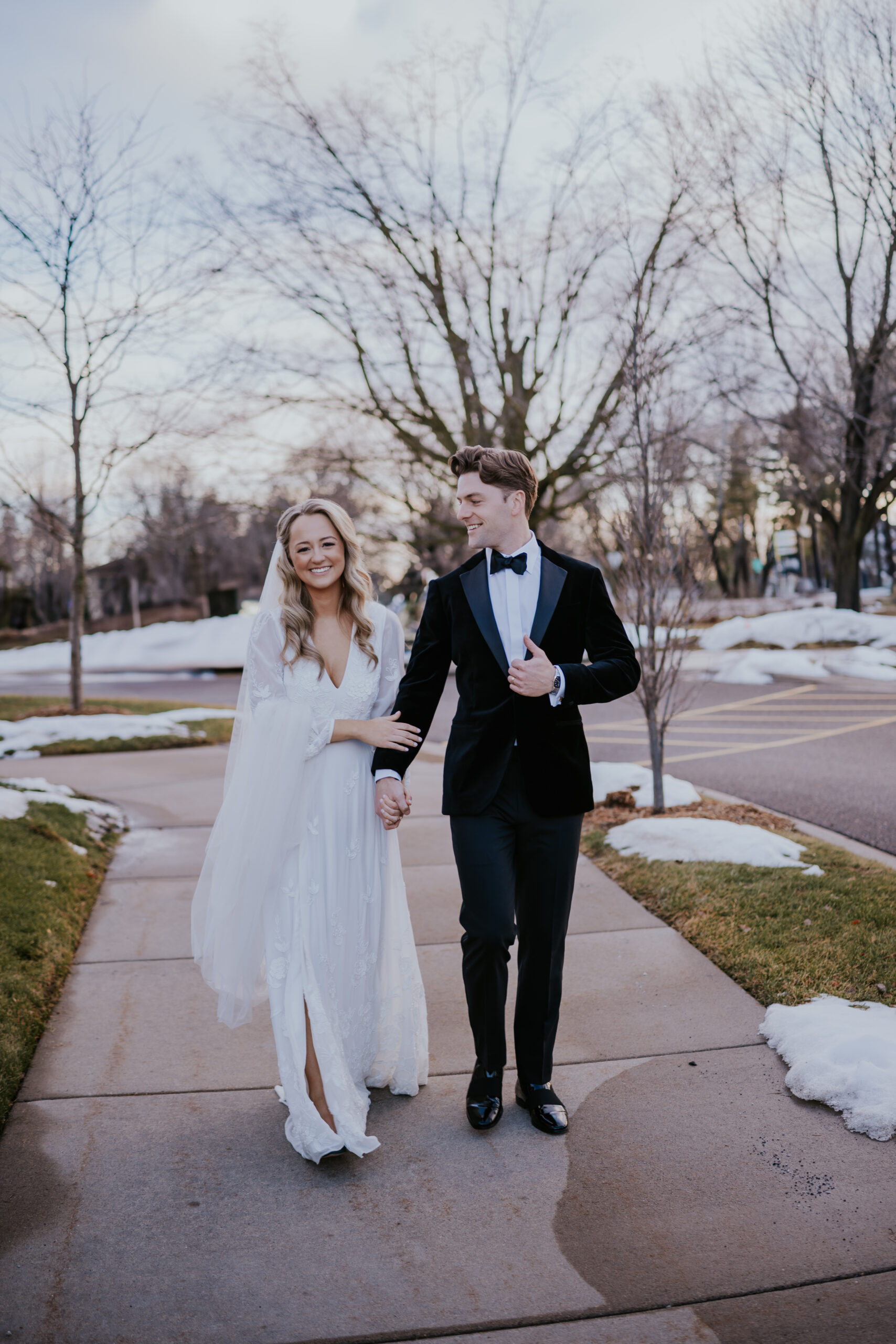 Bride and groom walking hand in hand down a snowy sidewalk, smiling at each other, photographed by a wedding photographer in Minnesota.