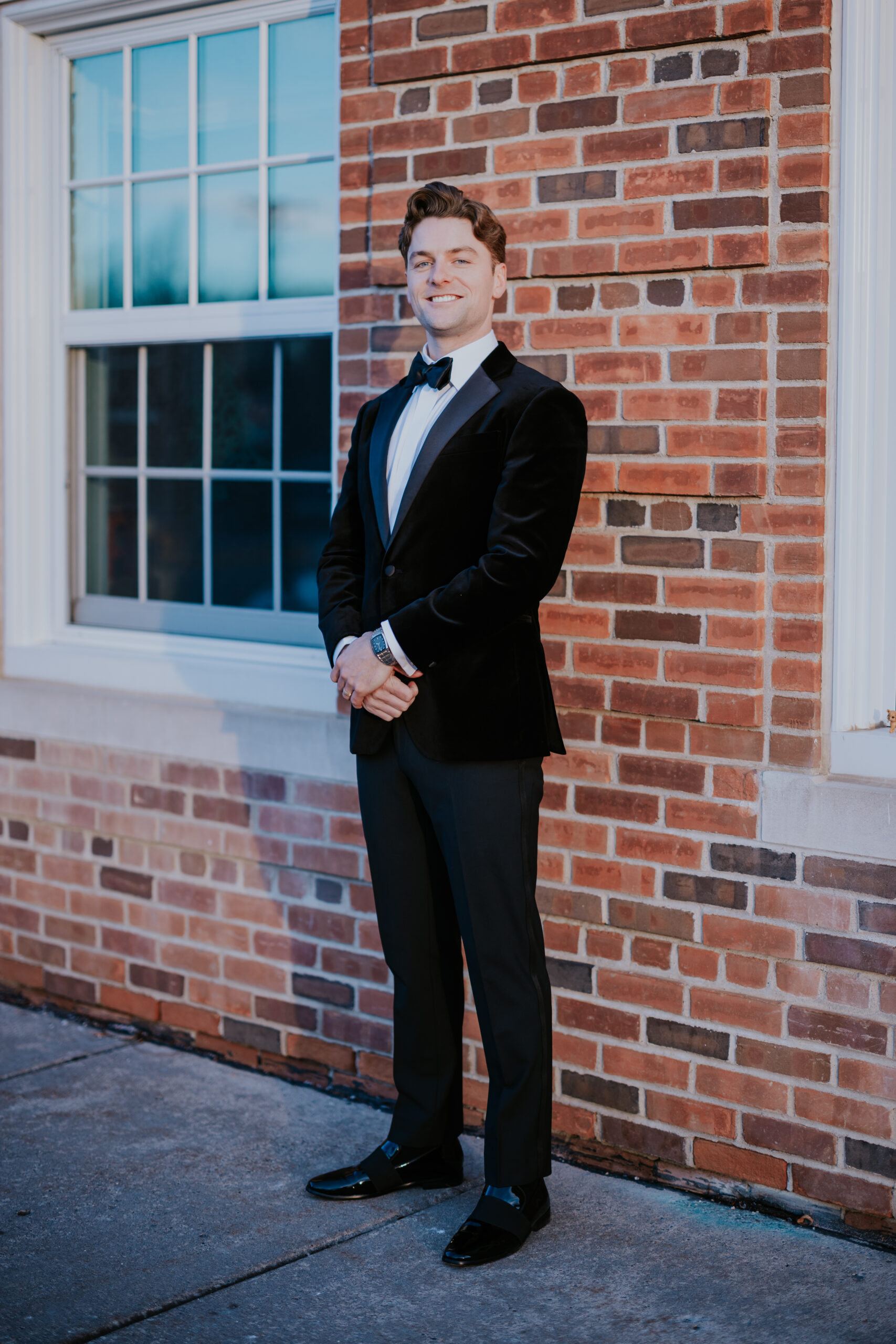 Groom posing confidently in a tuxedo against a brick wall with a window behind him.