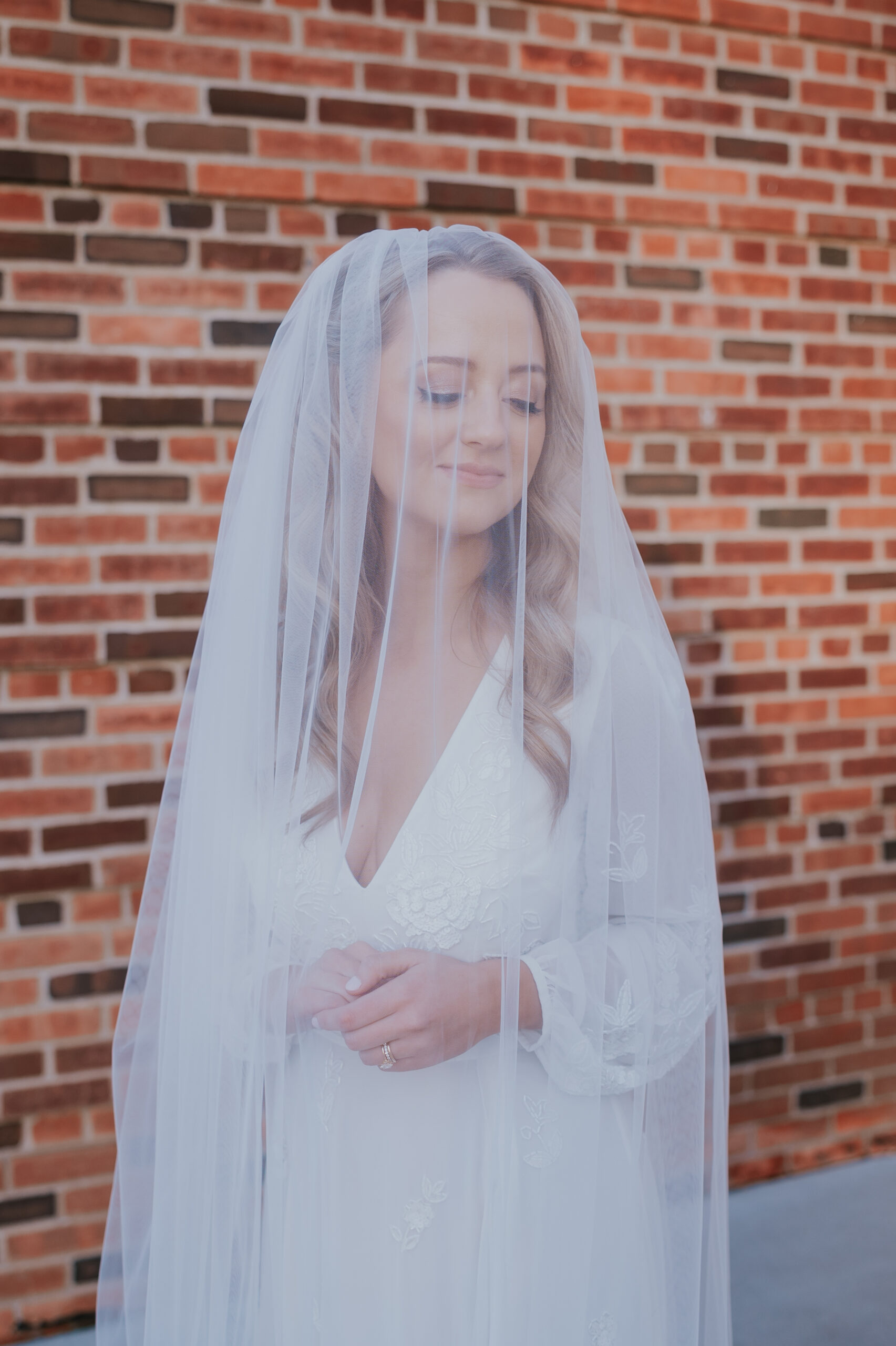 Bride with her veil draped over her face, softly smiling against a brick backdrop.