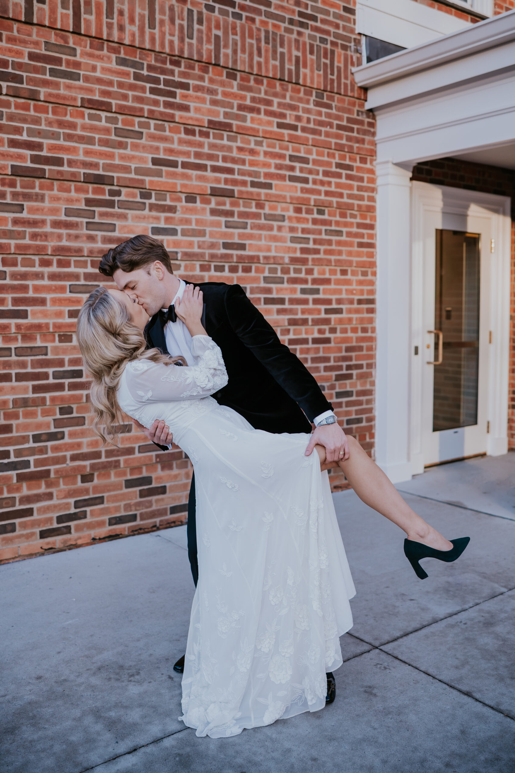 Groom dipping the bride for a kiss in front of a brick wall and white columns.