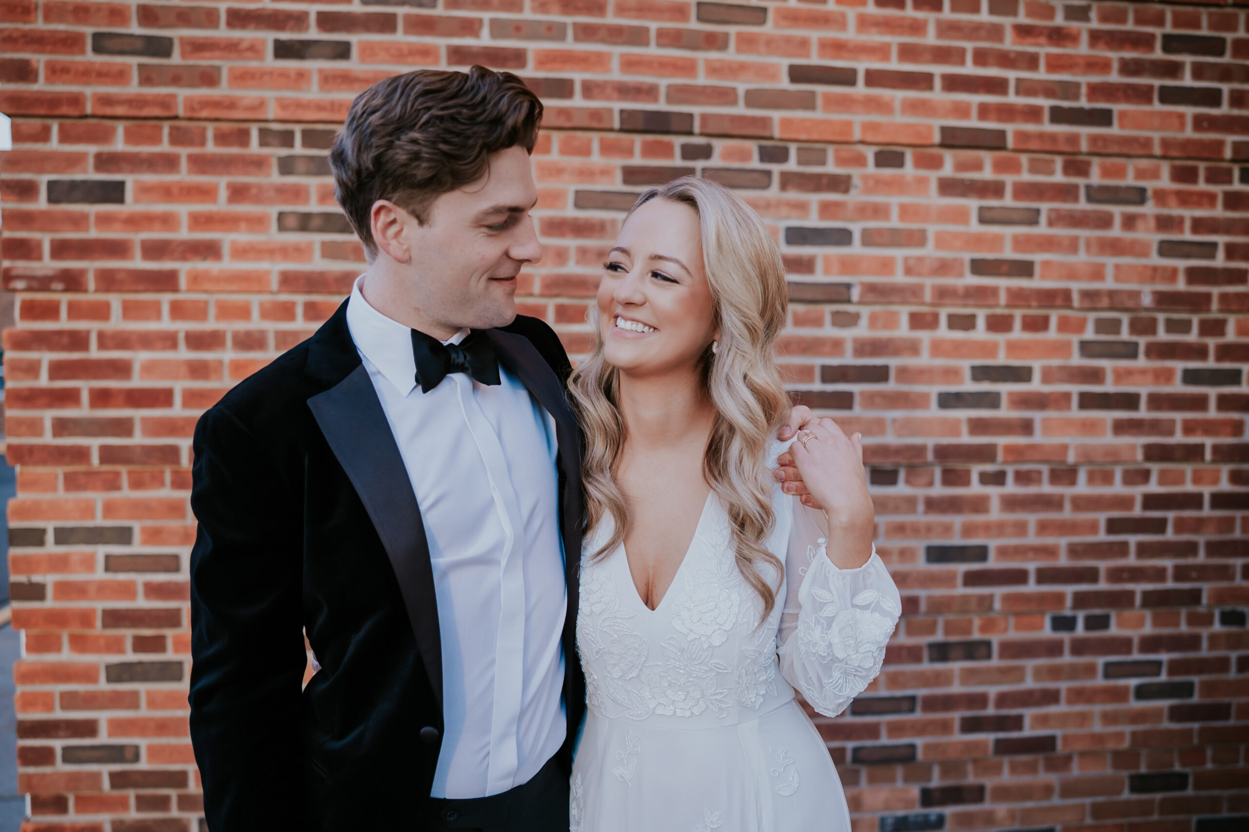 Bride and groom smiling at each other in front of a brick wall during their portraits.