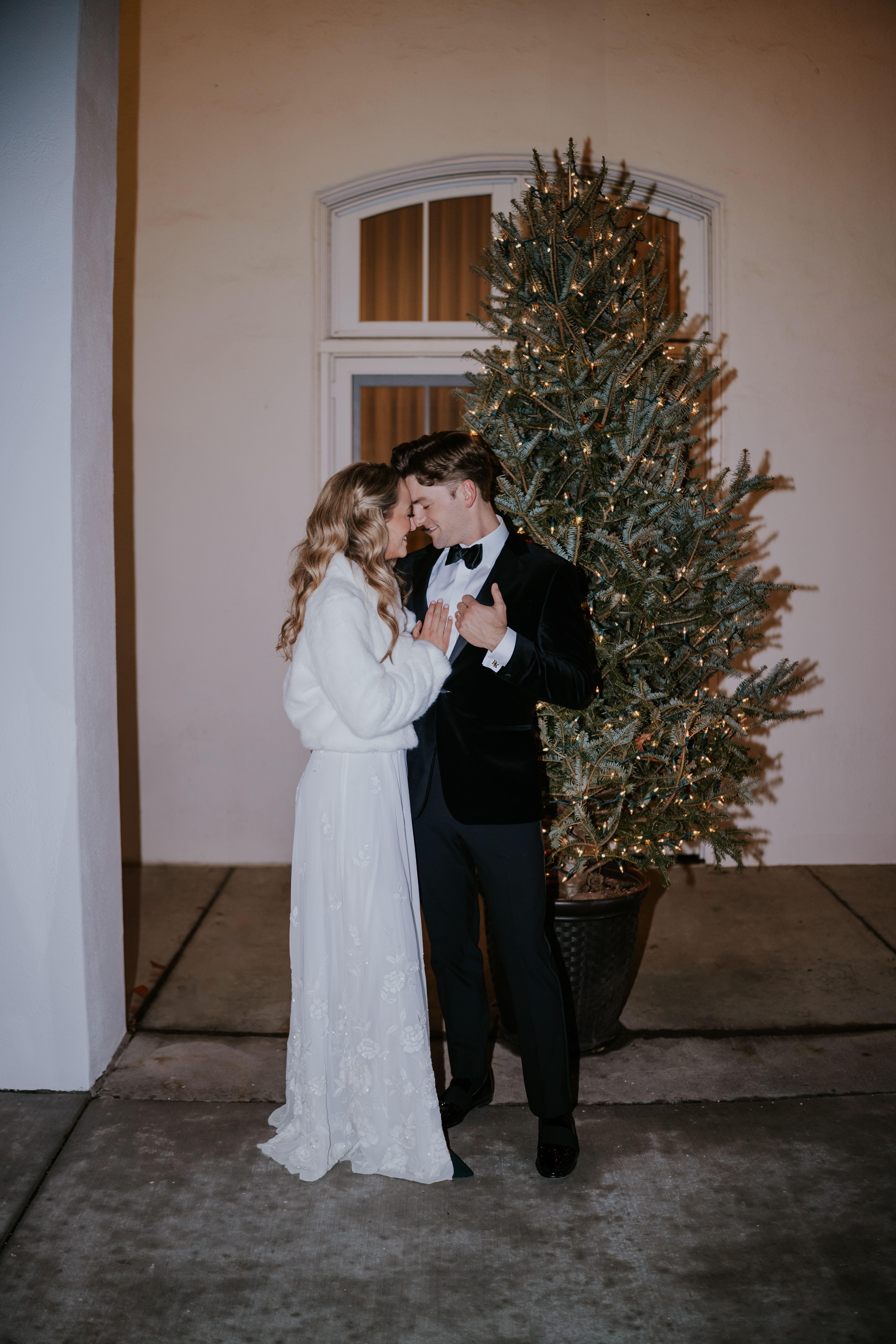 Couple holding each other closely in front of a Christmas tree decorated with twinkle lights.