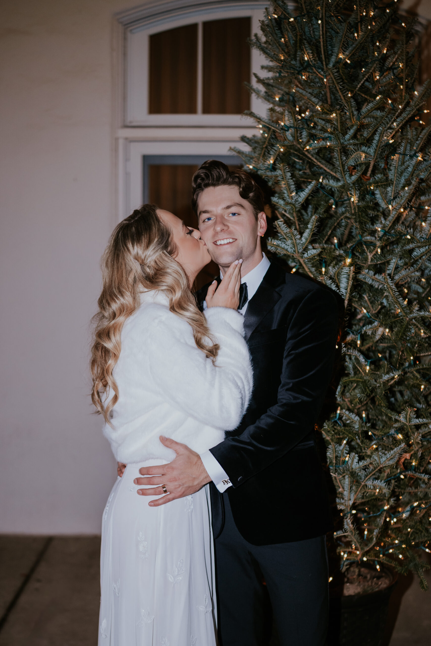 Bride kissing the groom’s cheek while standing beside a lit holiday tree, documented by a wedding photographer in Minnesota.