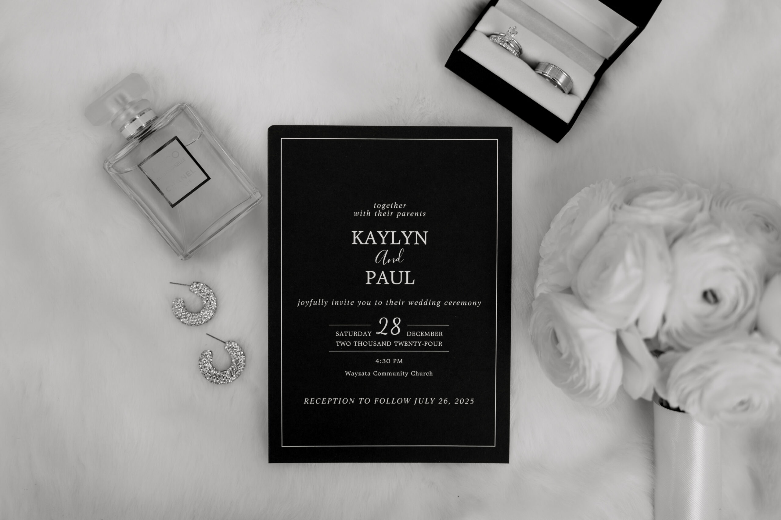 Black and white wedding invitation flat lay with perfume, earrings, rings, and bouquet.