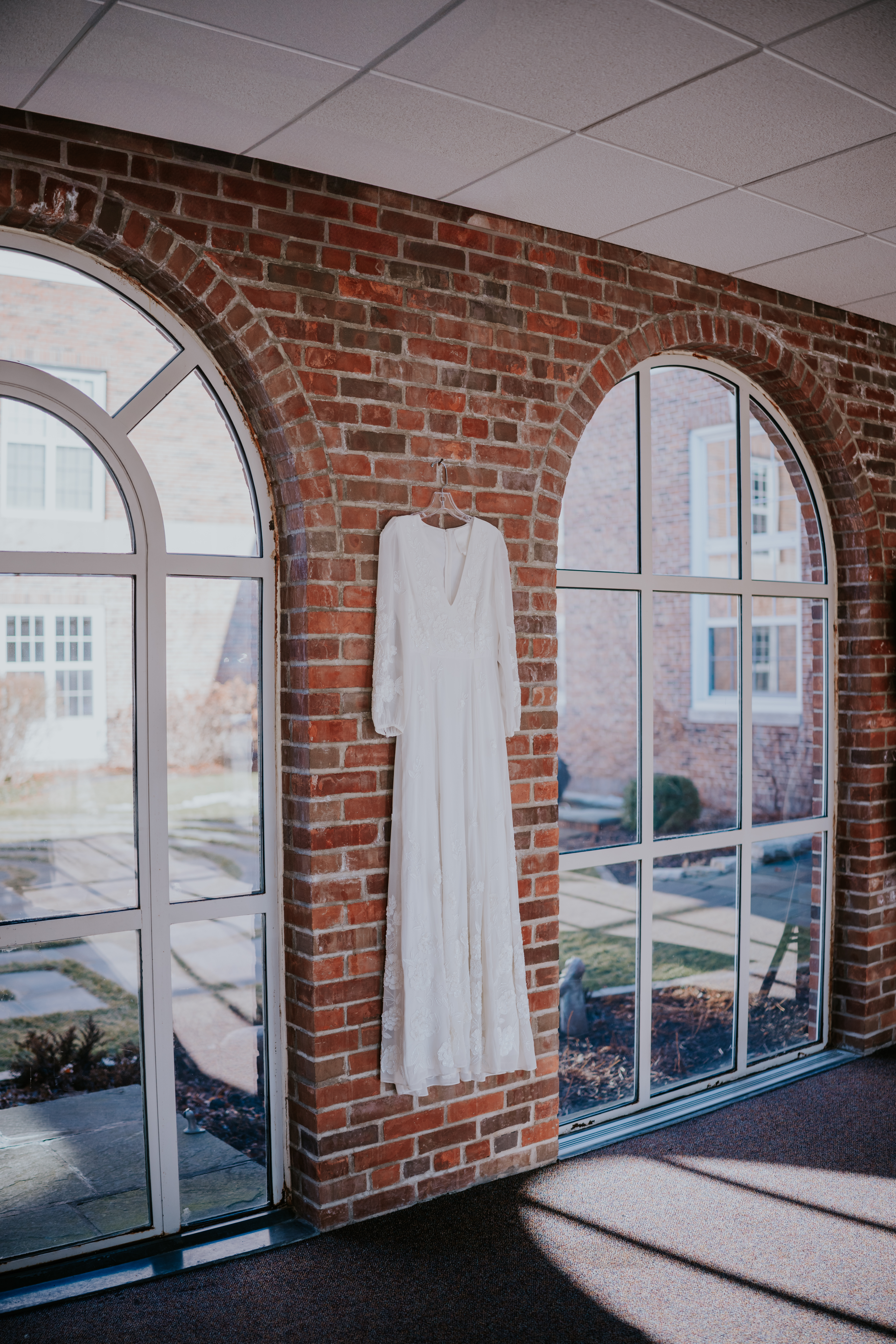Wedding dress hanging on a brick wall with tall arched windows, photographed by a wedding photographer in Minnesota.