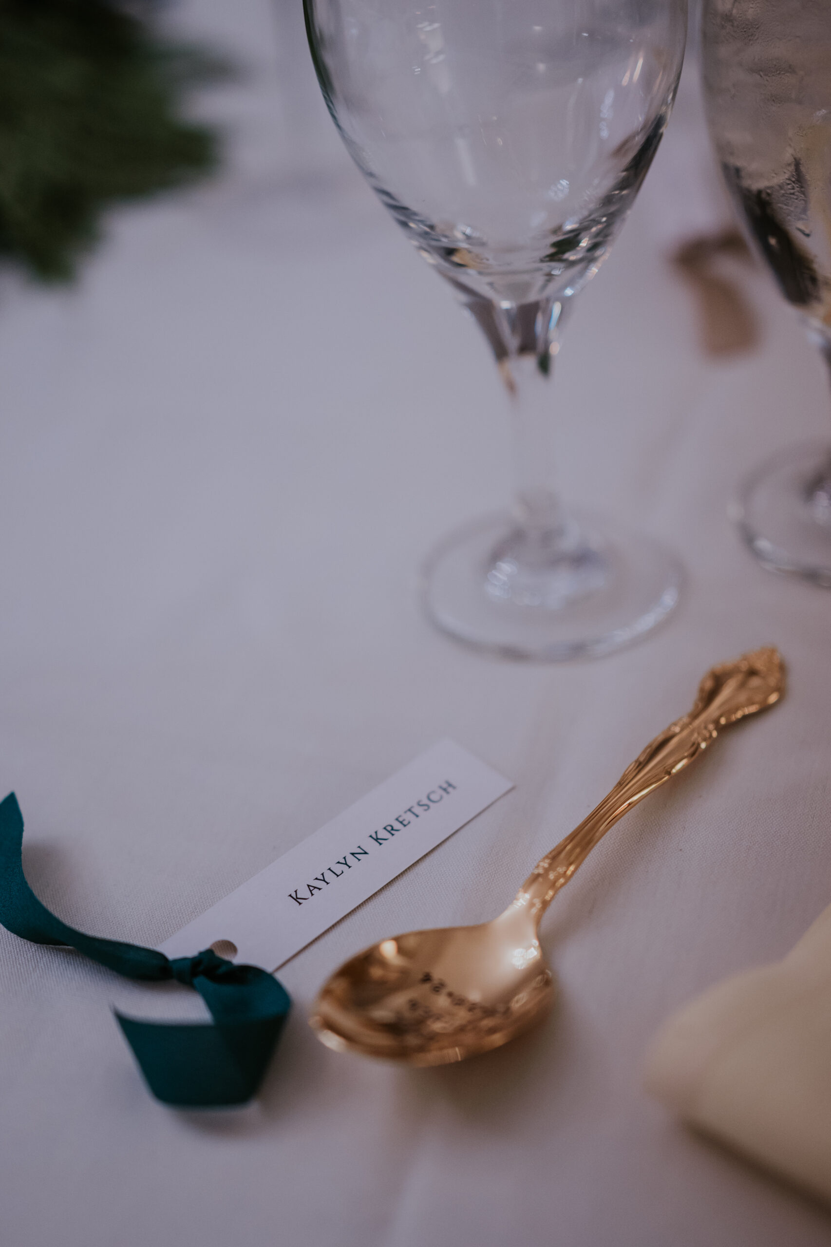 Close-up of a place setting with a gold spoon, name card, and wine glass on a white linen table.