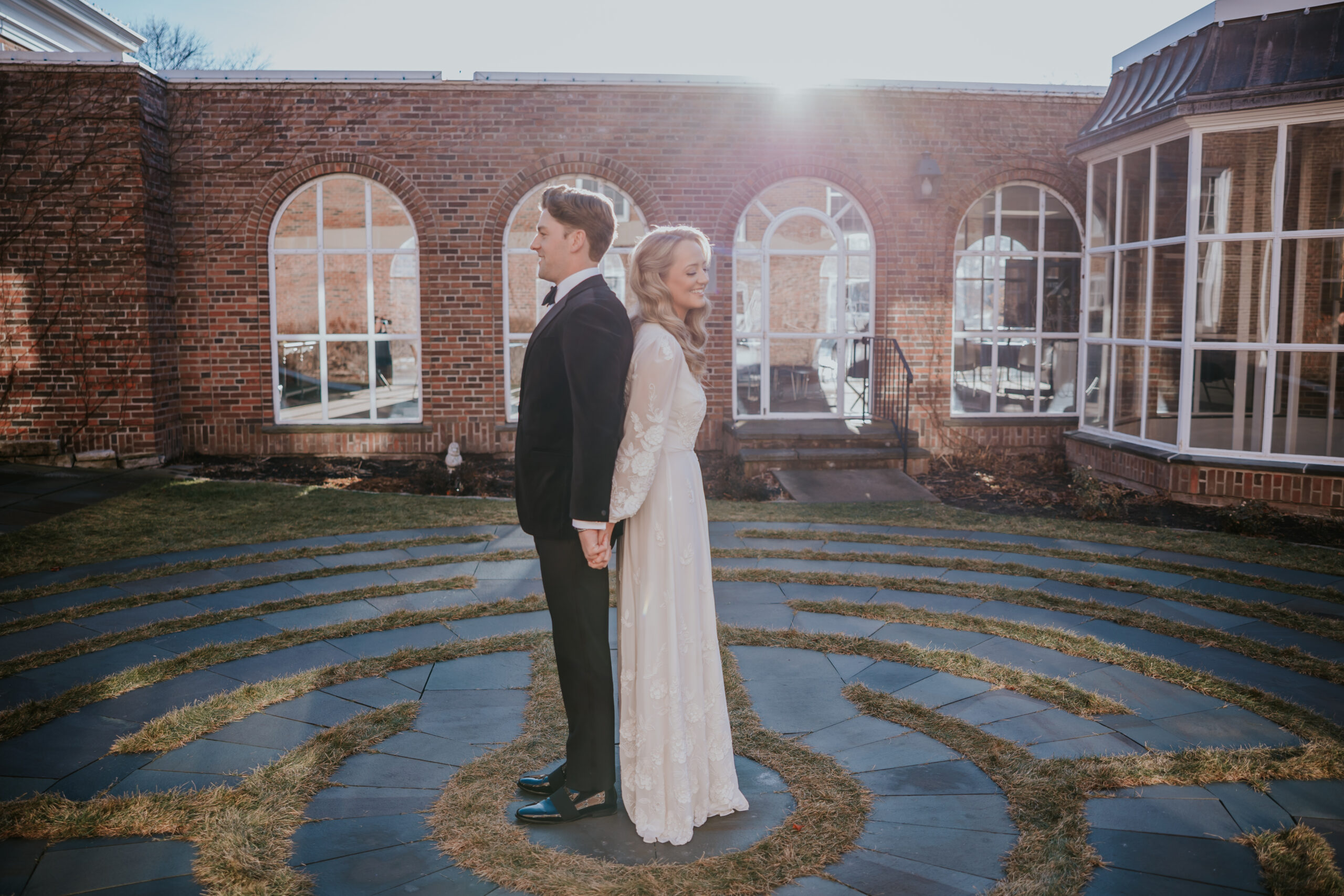 Bride and groom standing back to back, holding hands with the sun shining behind them.