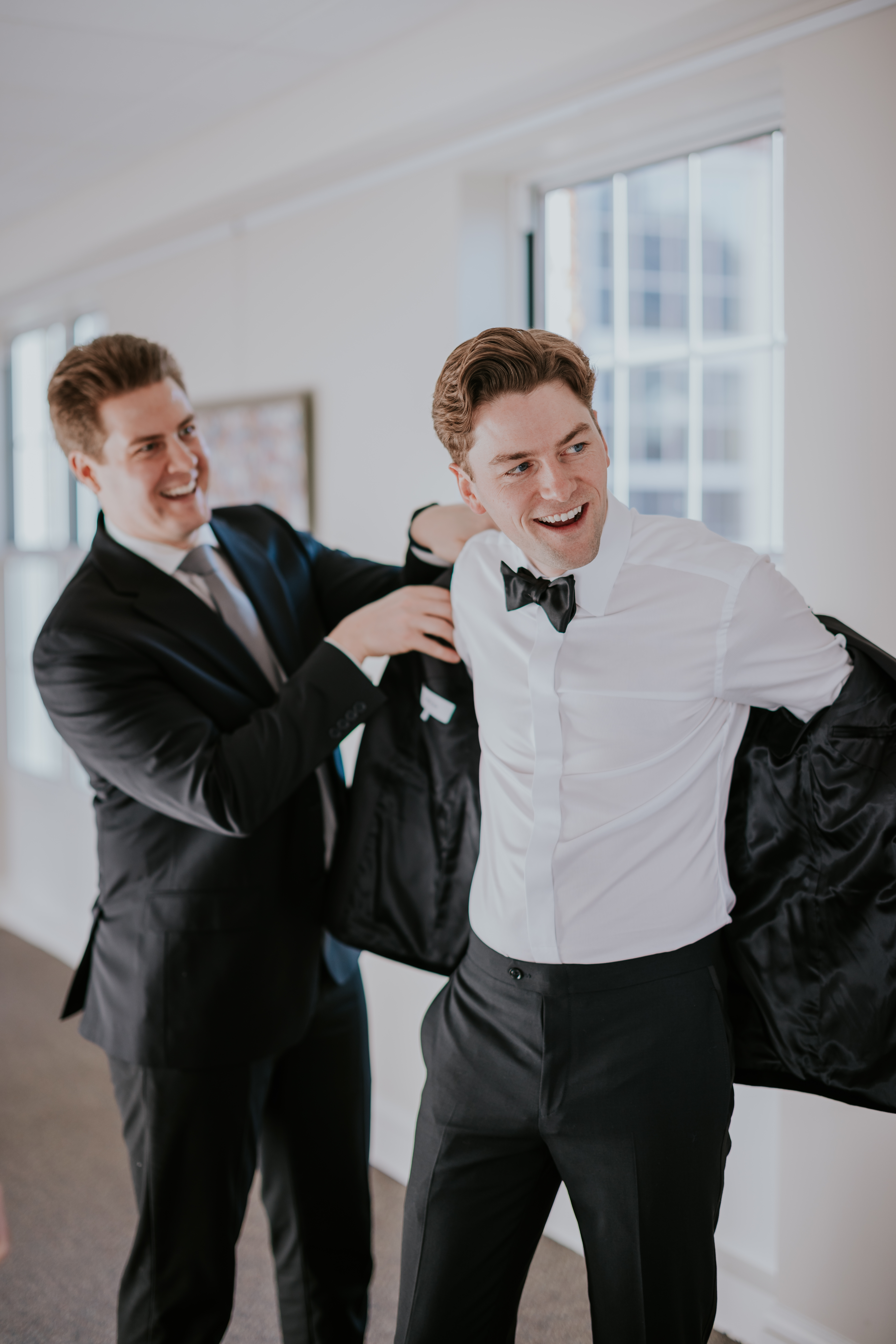 Groomsman helping the groom put on his jacket as he laughs, photographed by a wedding photographer in Minnesota.