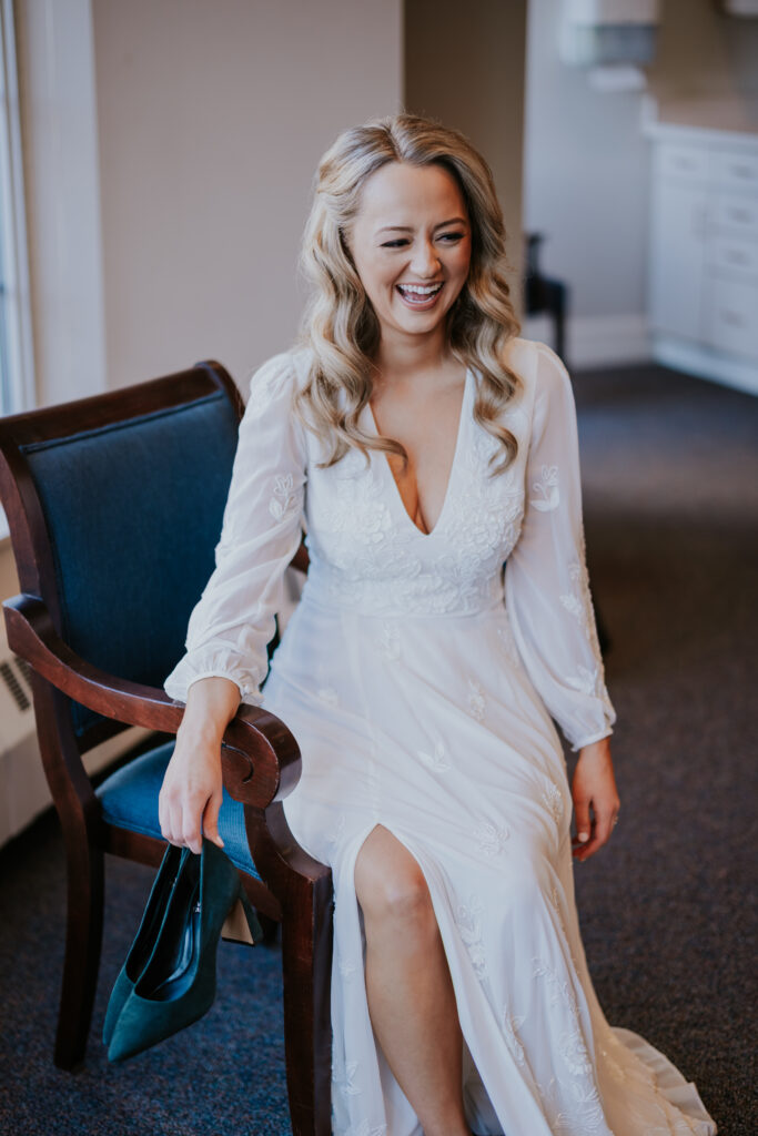 Bride laughing while sitting in a chair holding her shoes, captured by a wedding photographer in Minnesota.