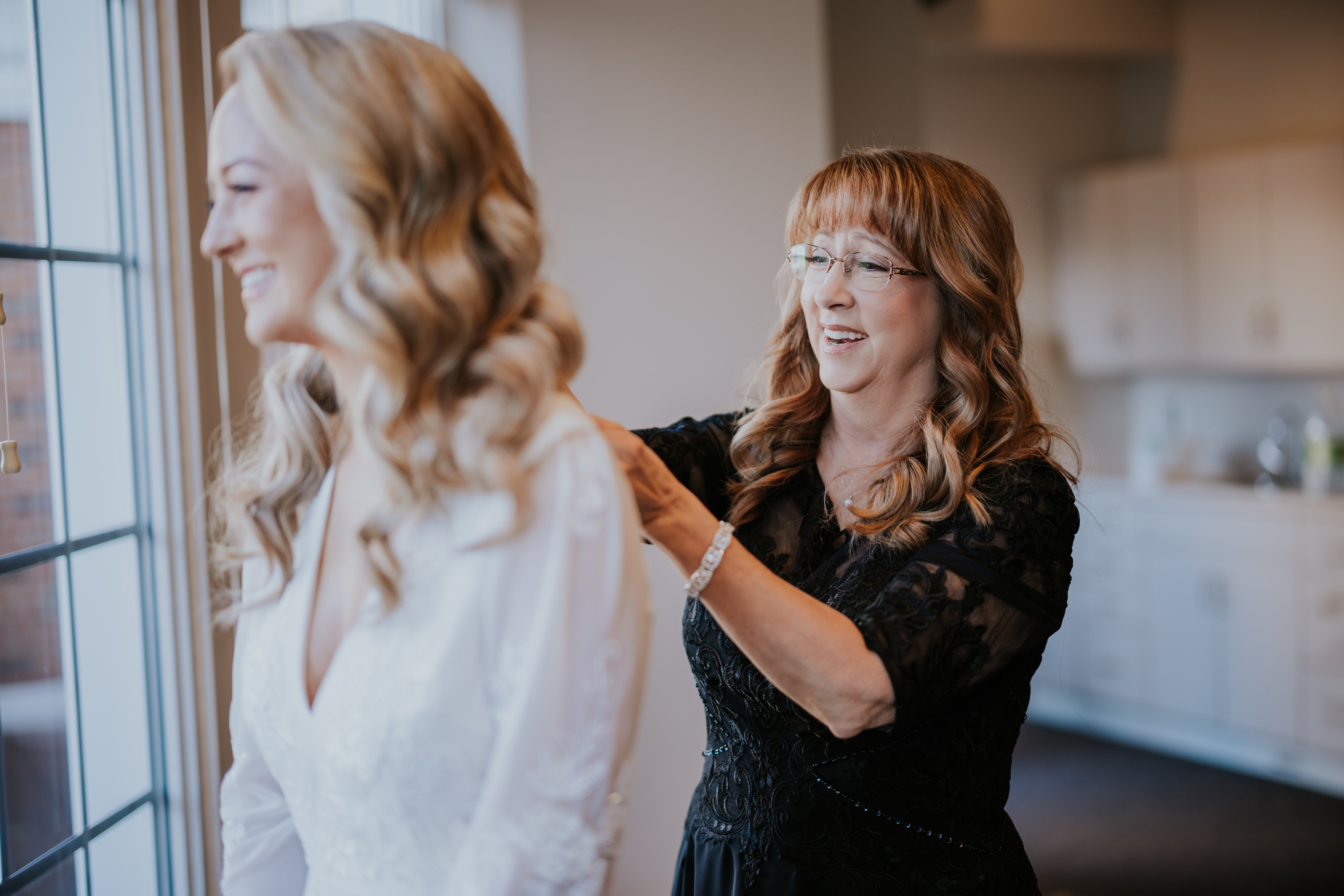 Mother of the bride fastening the back of the wedding dress while the bride smiles out the window.