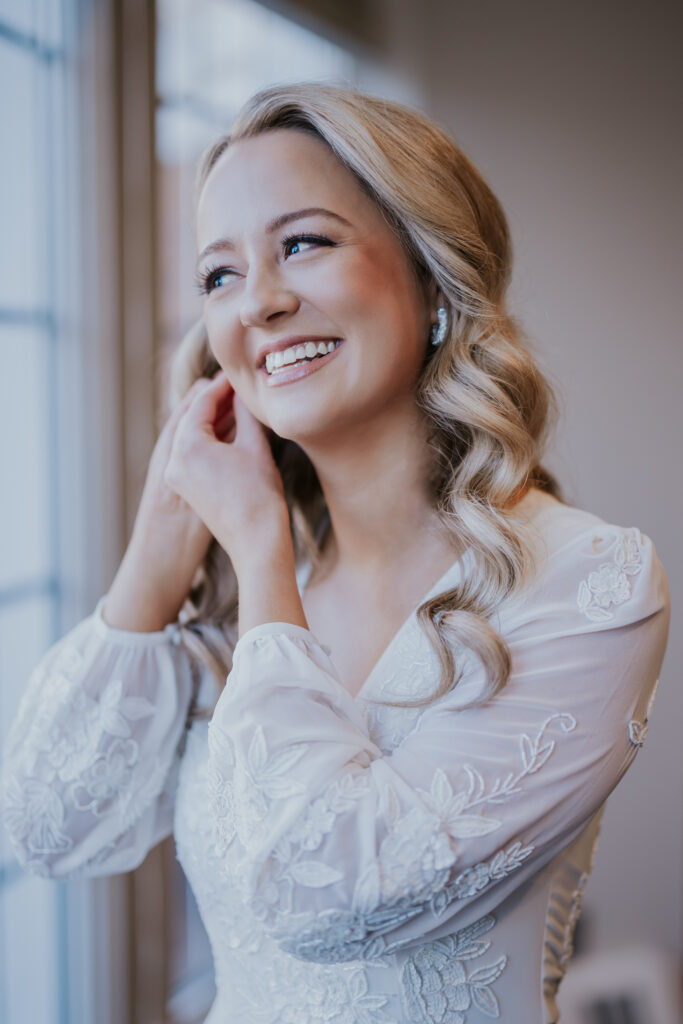 Bride smiling while adjusting her earrings near a window, photographed by a wedding photographer in Minnesota.