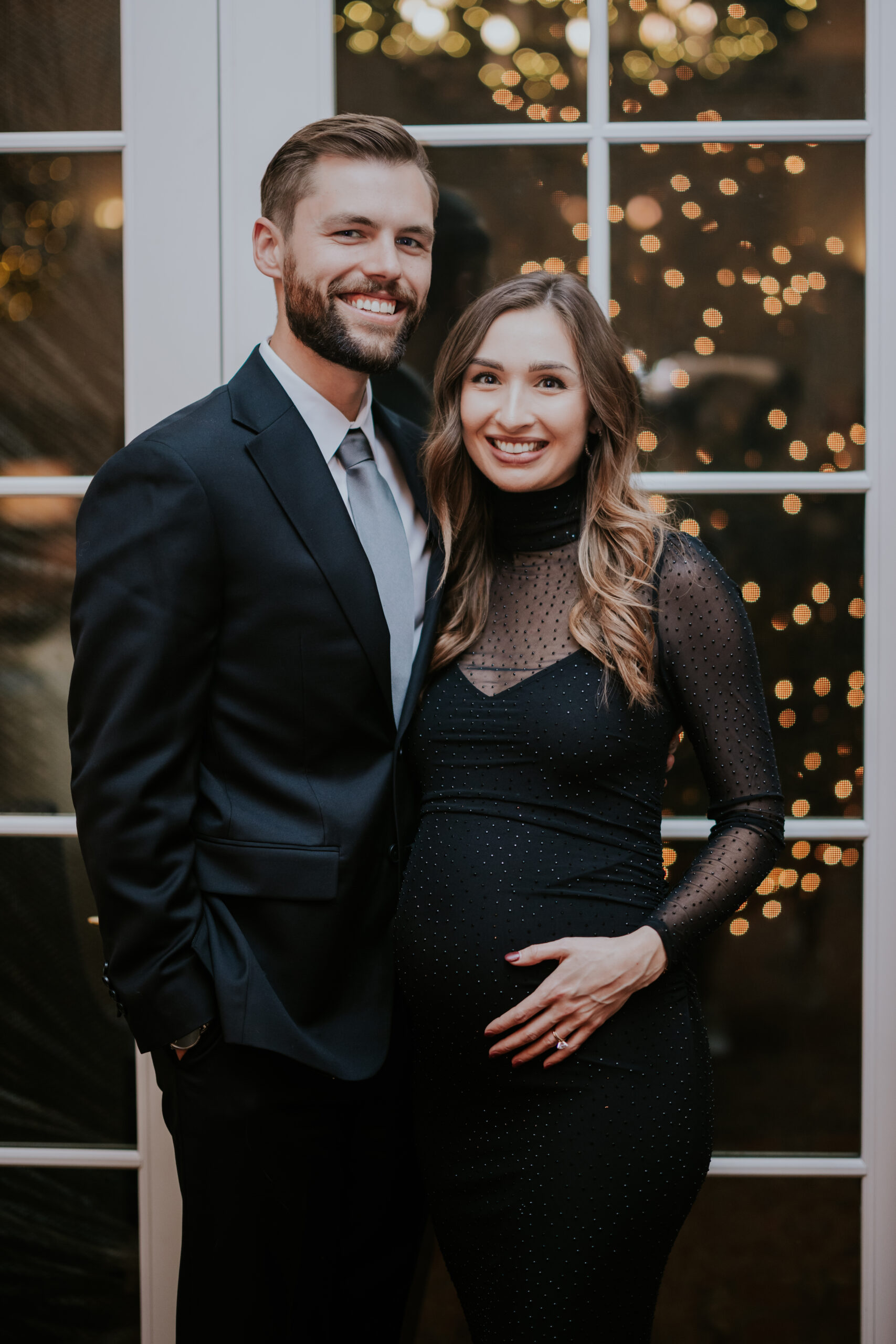 Couple posing together in front of glass doors with twinkle lights, the woman cradling her baby bump.