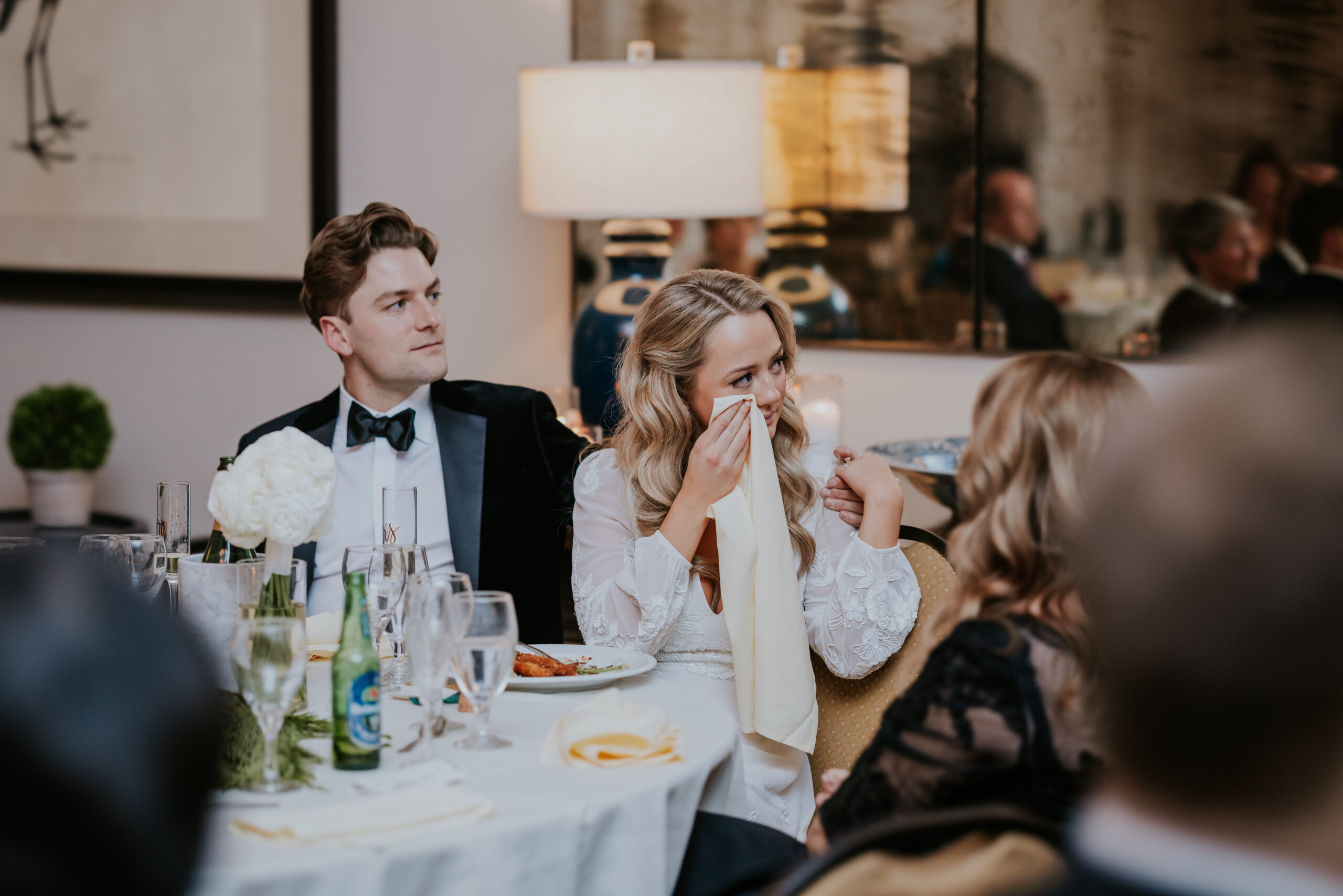 Bride wiping away tears at the reception while holding hands with a guest, groom sitting beside her.