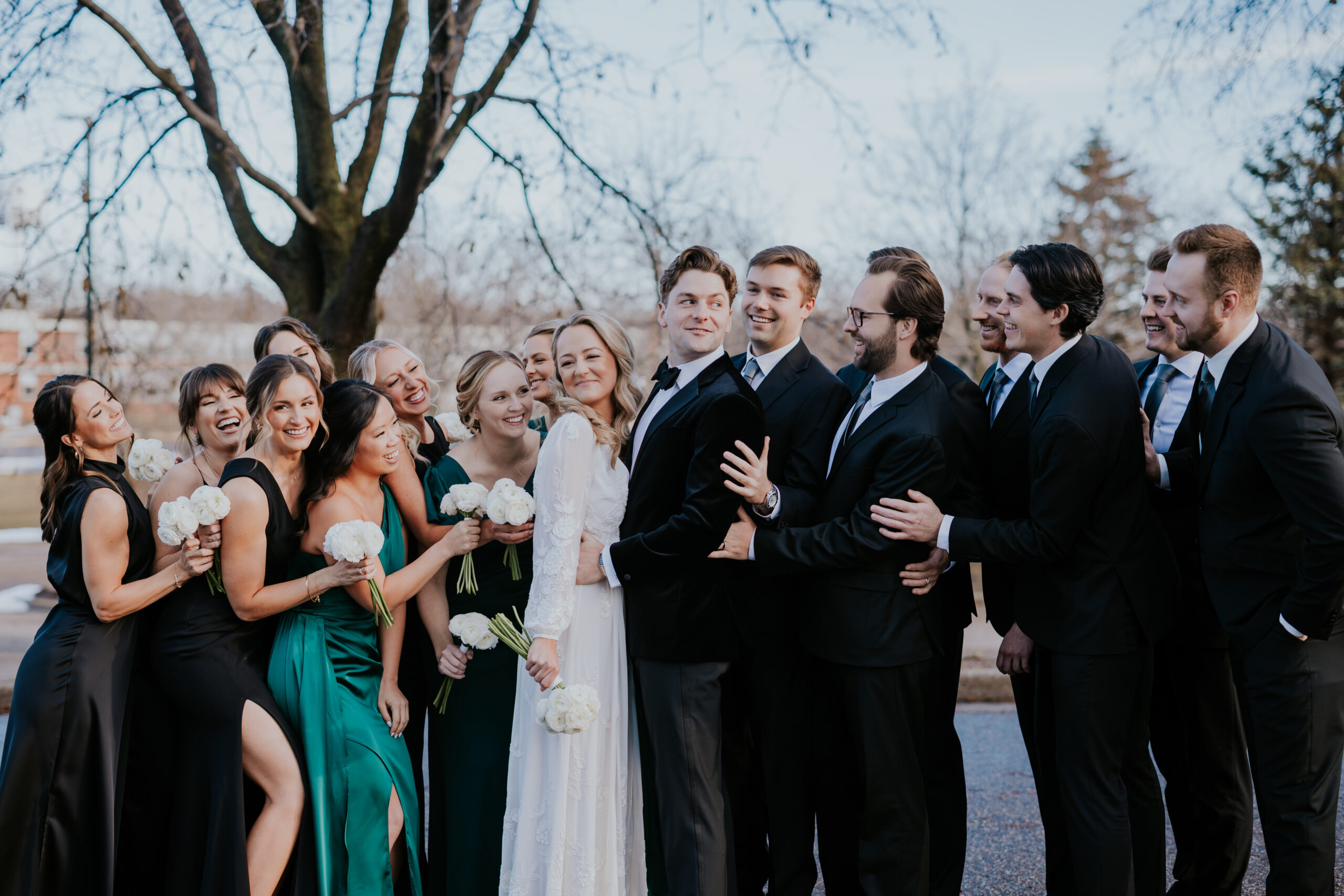 Large wedding party gathered around the bride and groom outdoors, everyone laughing and hugging, photographed by a wedding photographer in Minnesota.