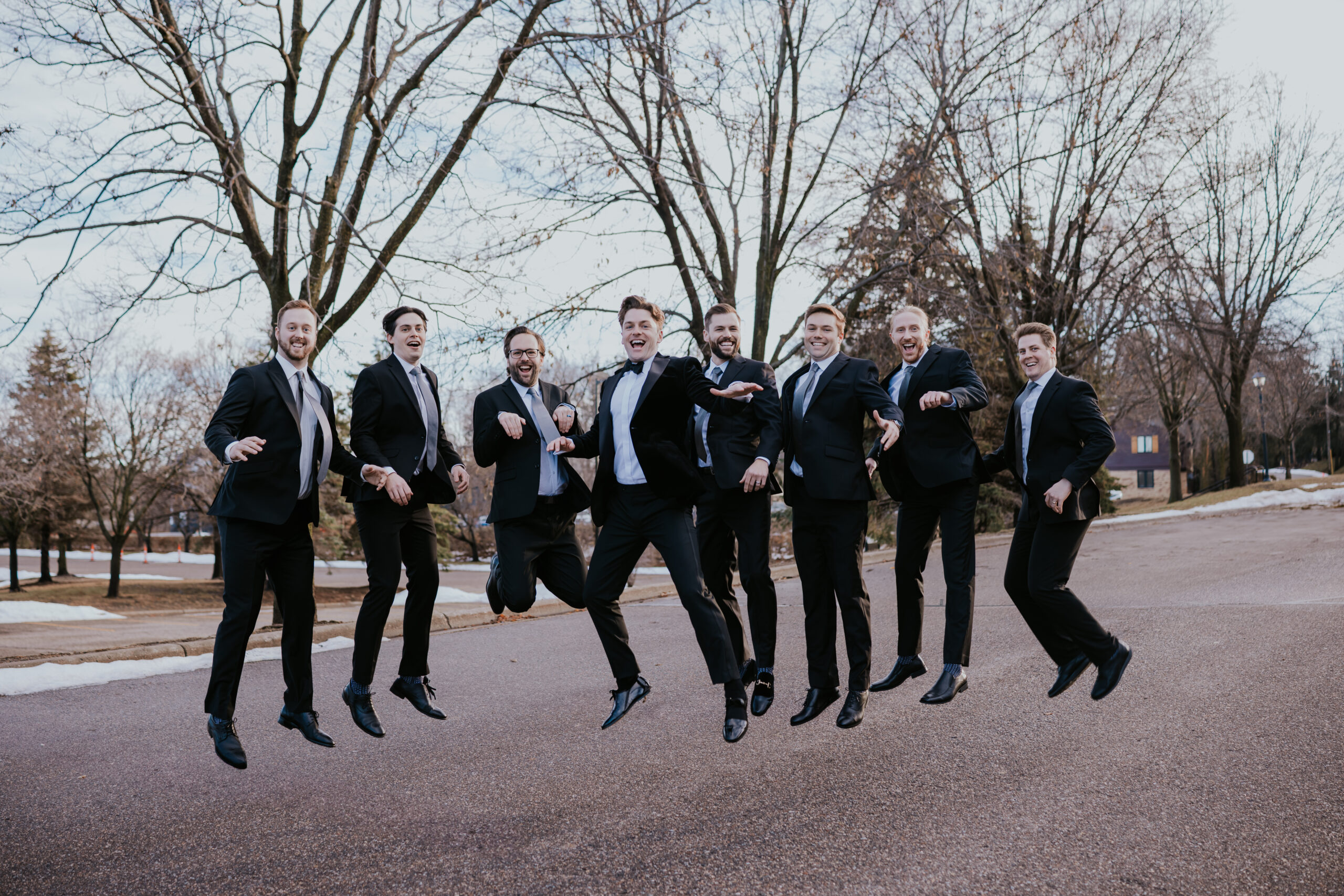 Groomsmen in black tuxedos jumping in the air together on a street.