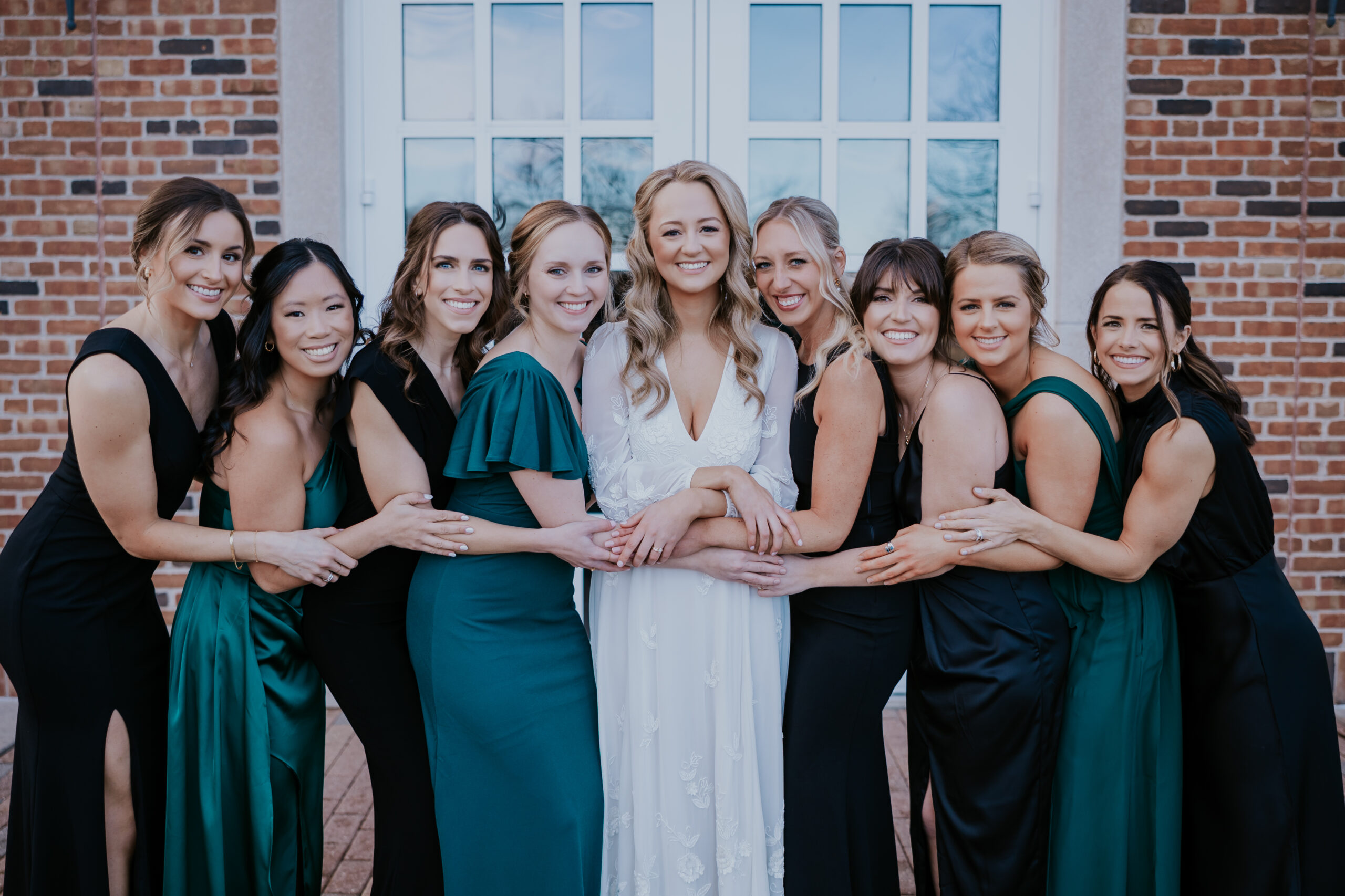 Bride surrounded by bridesmaids in green and black gowns, all smiling and holding her hands.