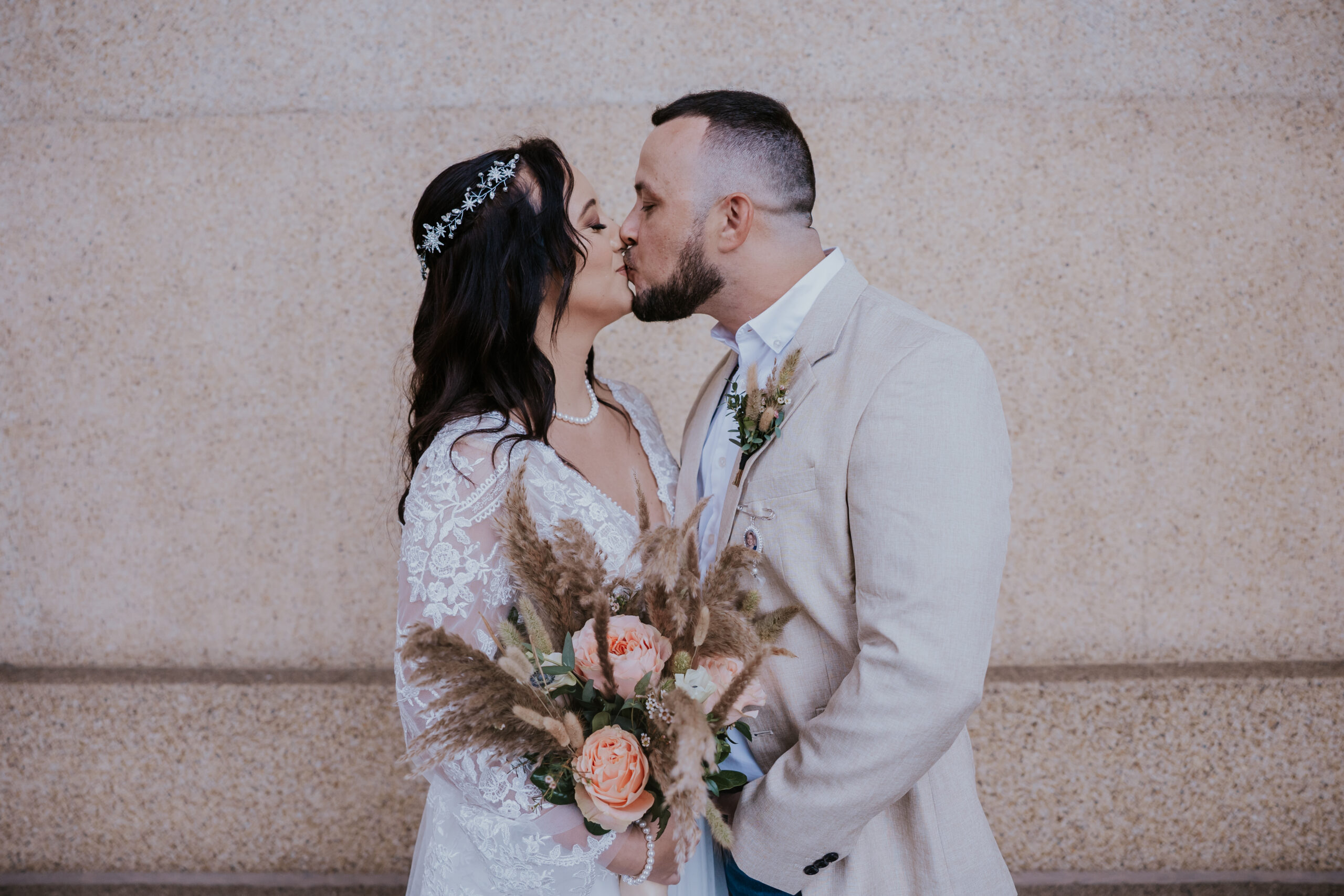 Bride and groom kiss in front of a textured stone wall, holding a boho bouquet of peach garden roses and pampas grass.