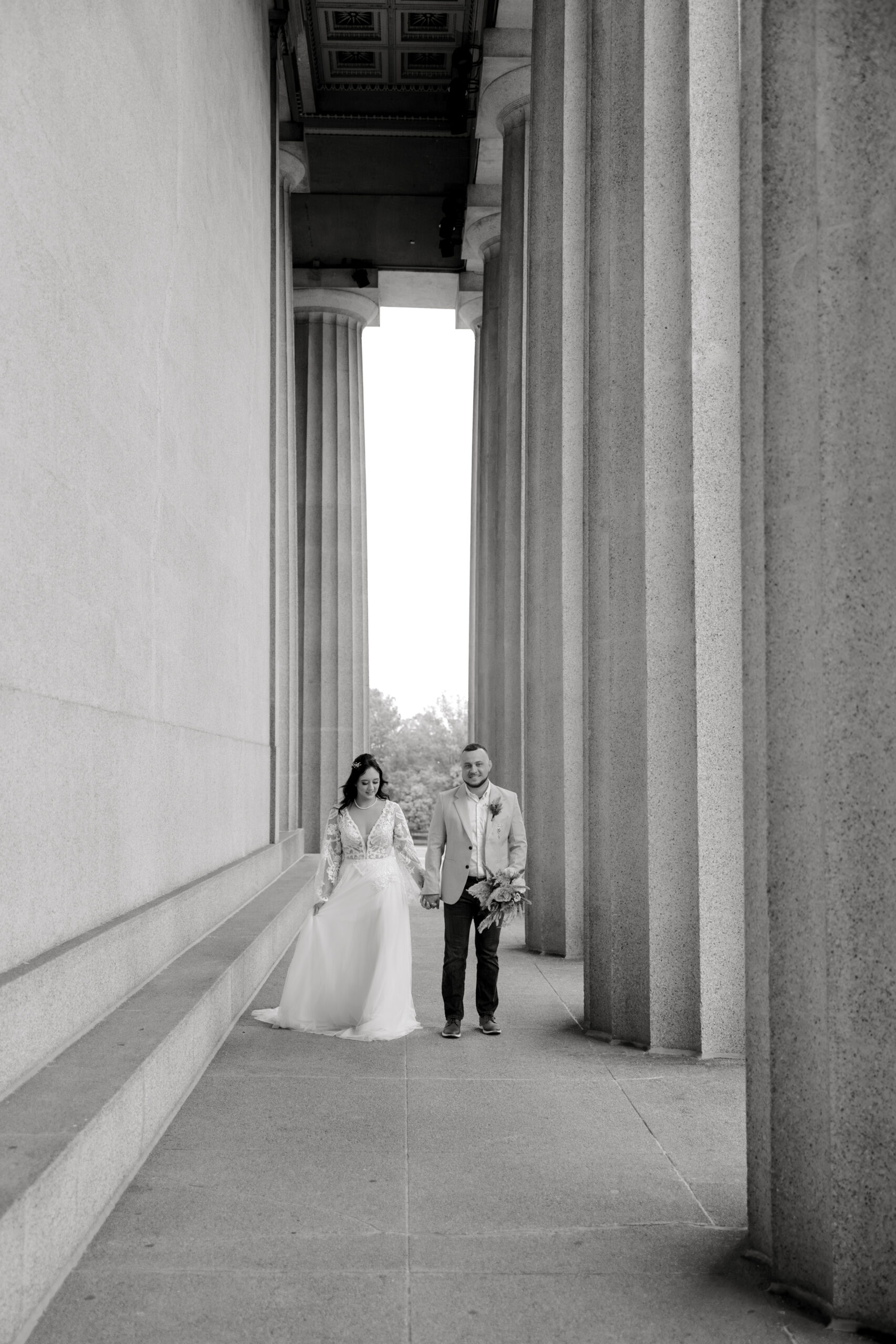 Bride and groom walk hand in hand down a column-lined walkway, a classic black and white image by a travel wedding photographer.