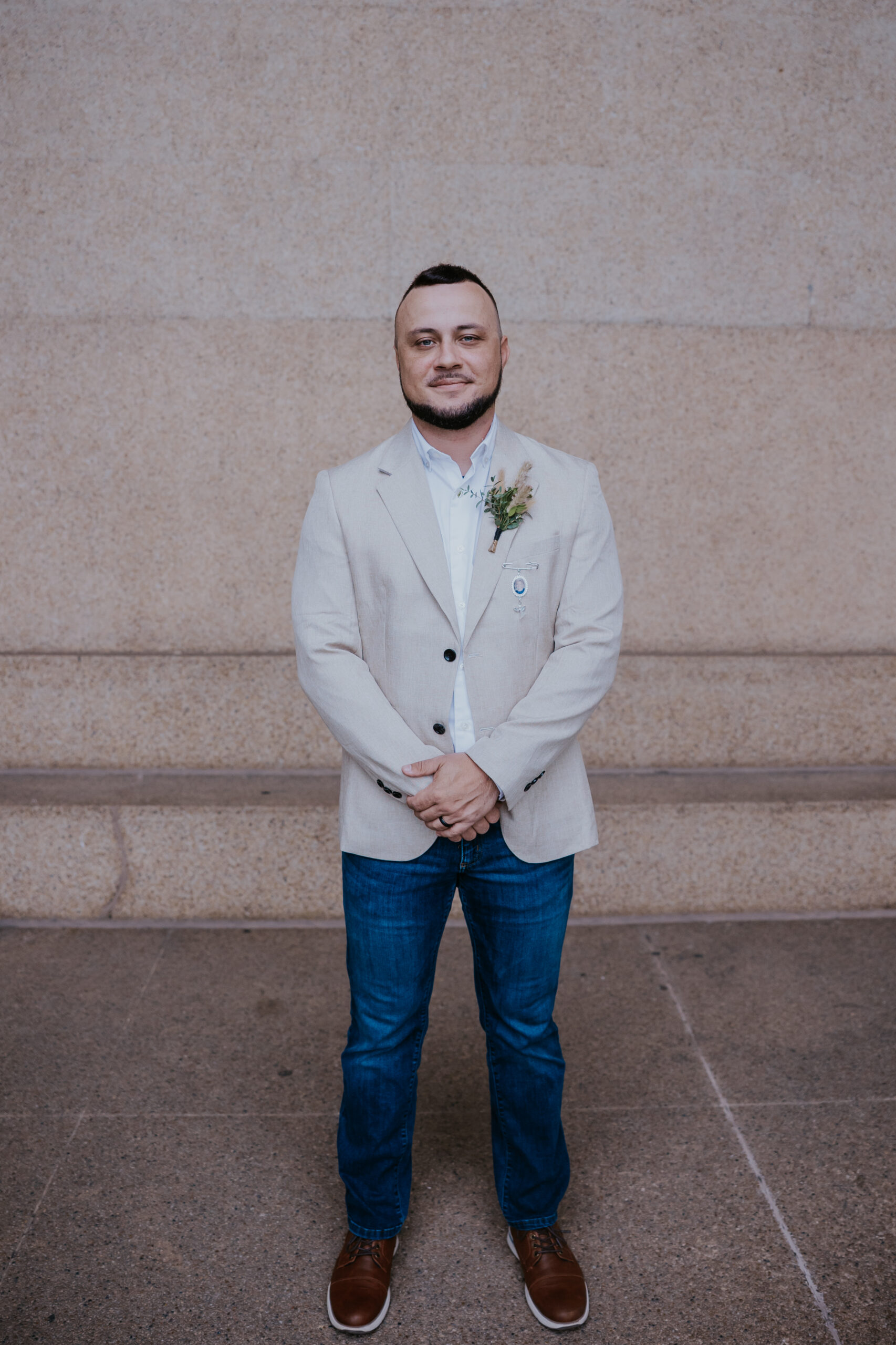 Groom posing for a wedding portrait in a tan blazer and jeans, with a boutonniere and a relaxed, stylish vibe.